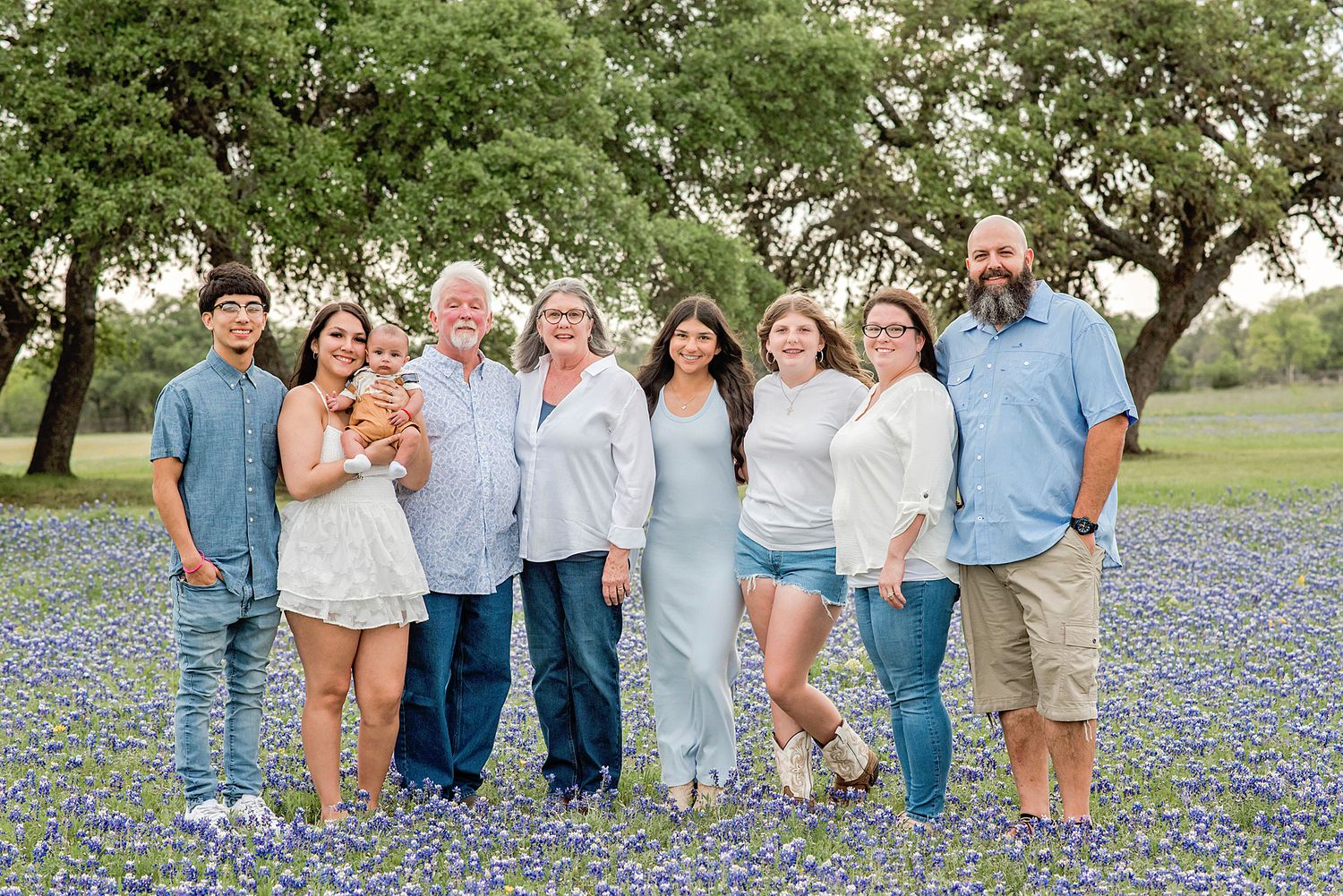 Family of eight smiling together in a Texas bluebonnet field surrounded by green trees on a sunny day.