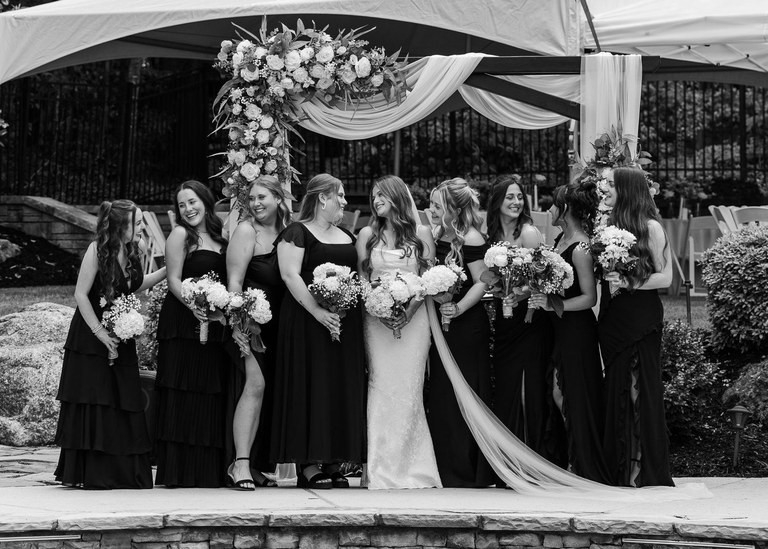 Black and white wedding photo of bride in white gown with bridesmaids in black dresses holding bouquets outdoors.
