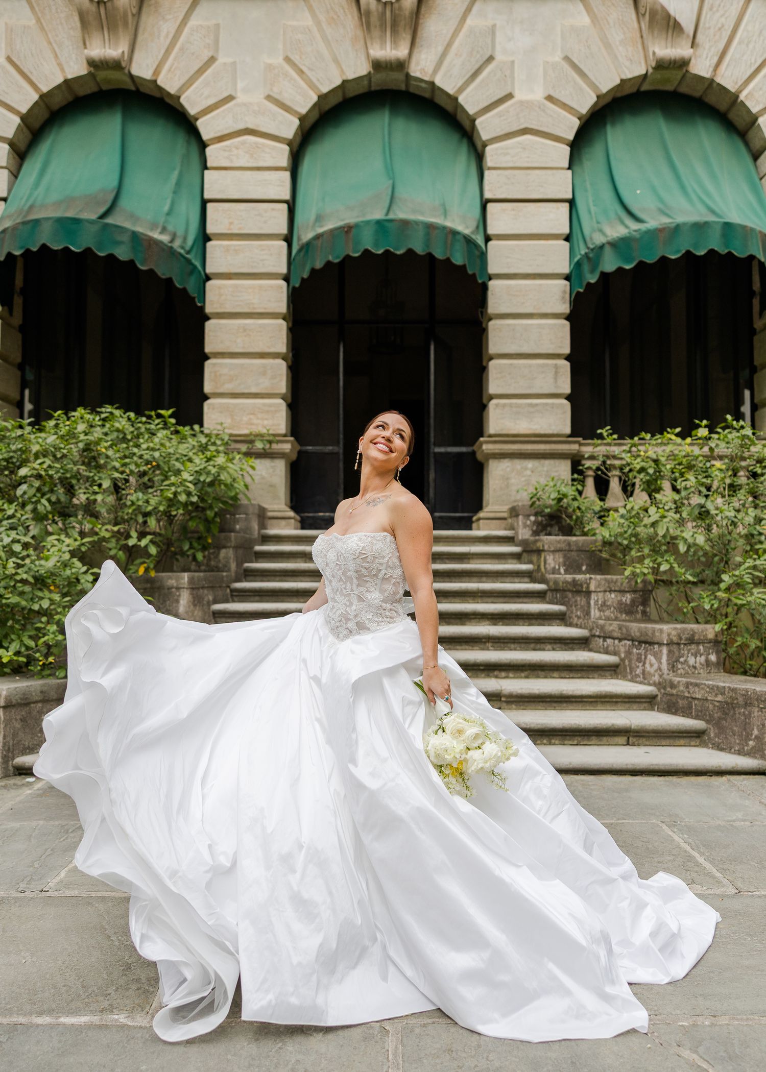 Bride in strapless white ballgown twirling outside elegant stone building with green awnings.