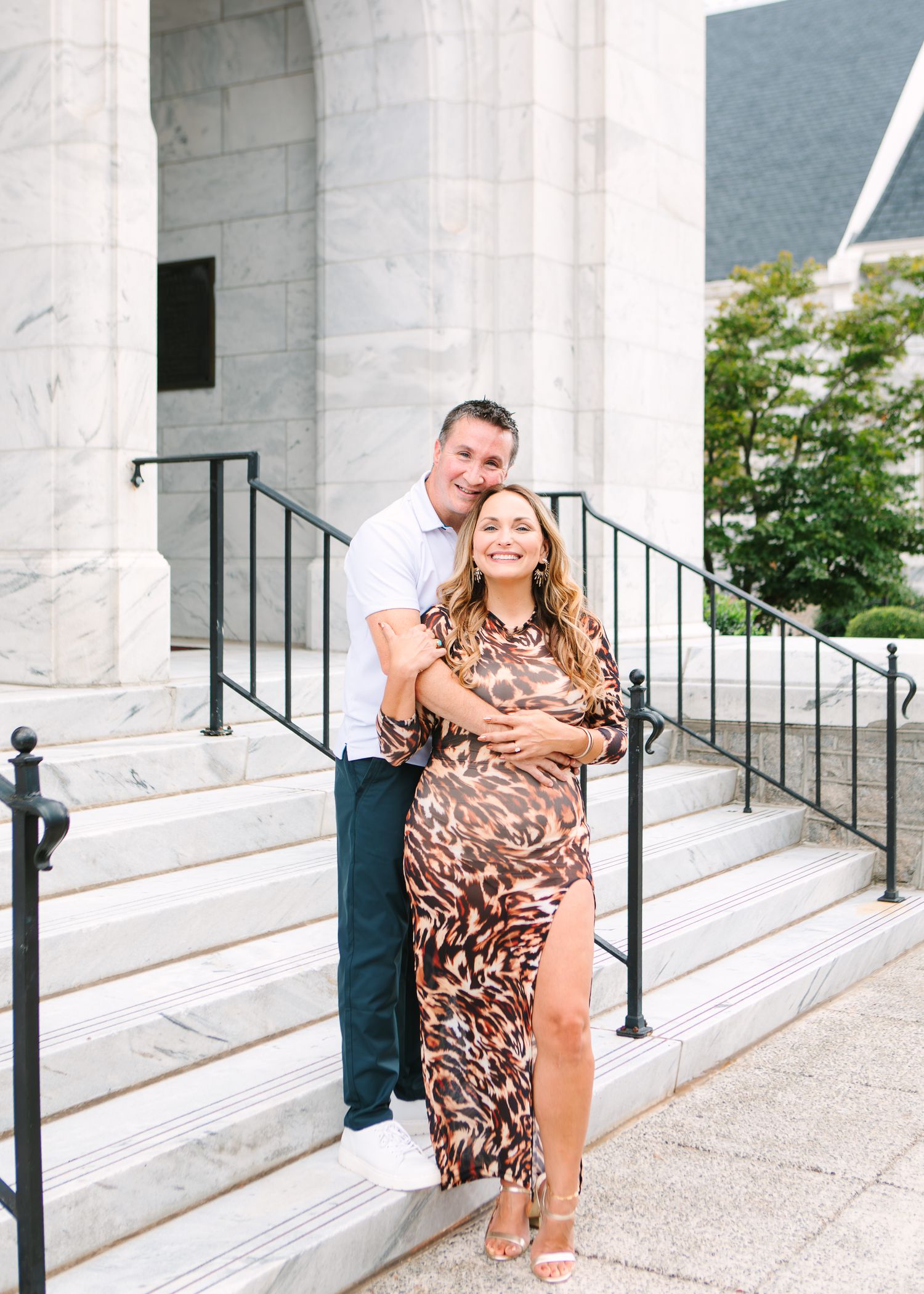 Couple posing on marble steps, woman in leopard print dress, man in white shirt, embracing outdoors.
