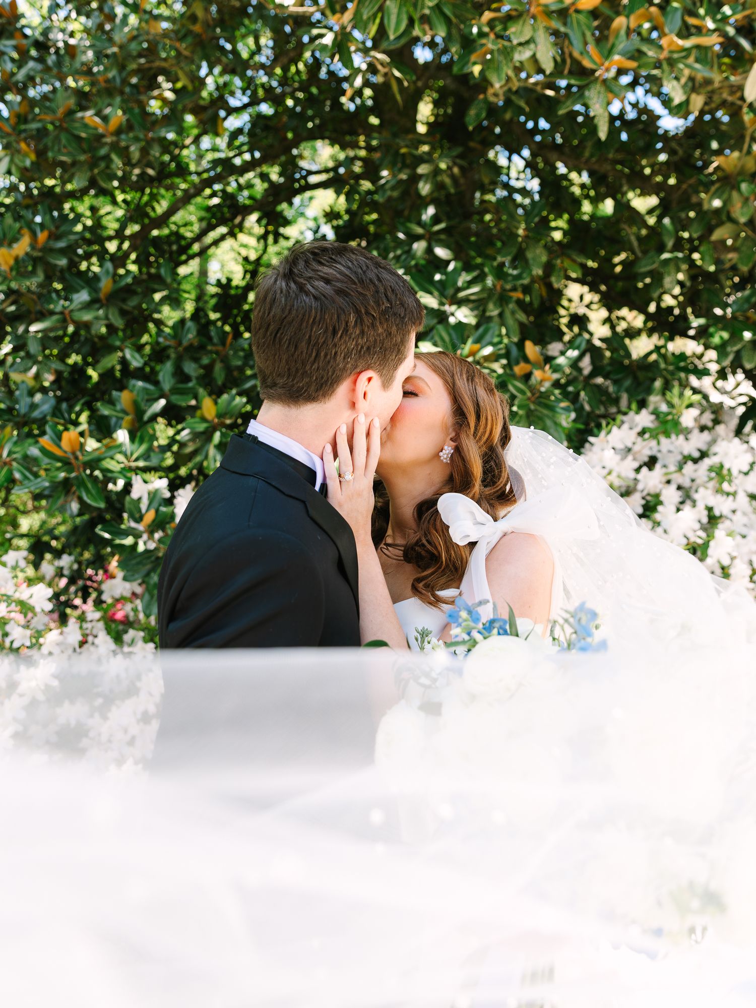 A romantic wedding moment captured under flowering trees with soft white veil details.