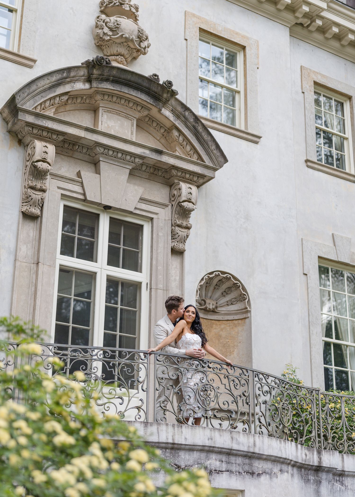 Bride and groom share a romantic moment on an ornate balcony of an elegant historic mansion.