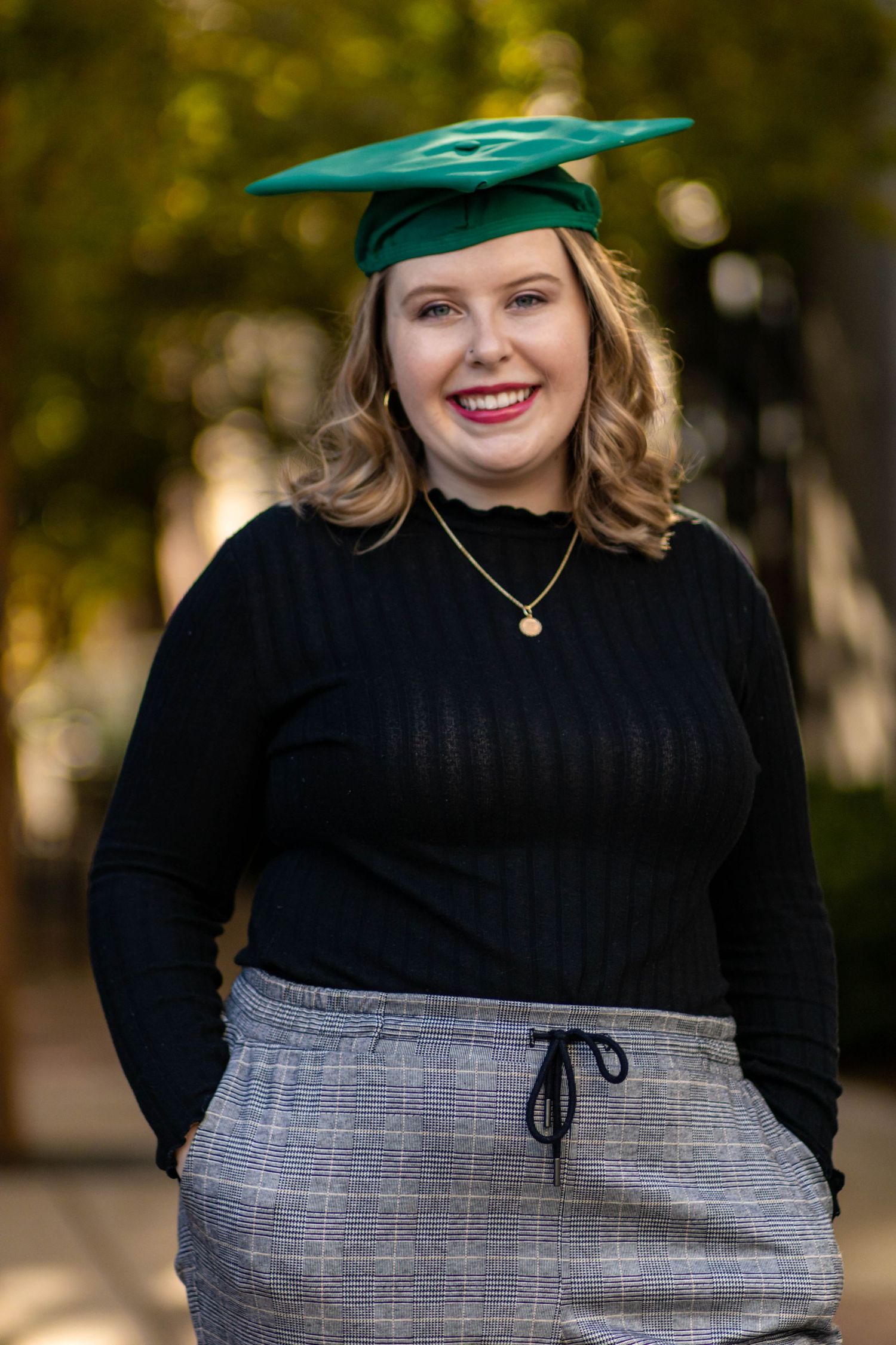 Graduate in black turtleneck and green academic regalia poses outdoors.