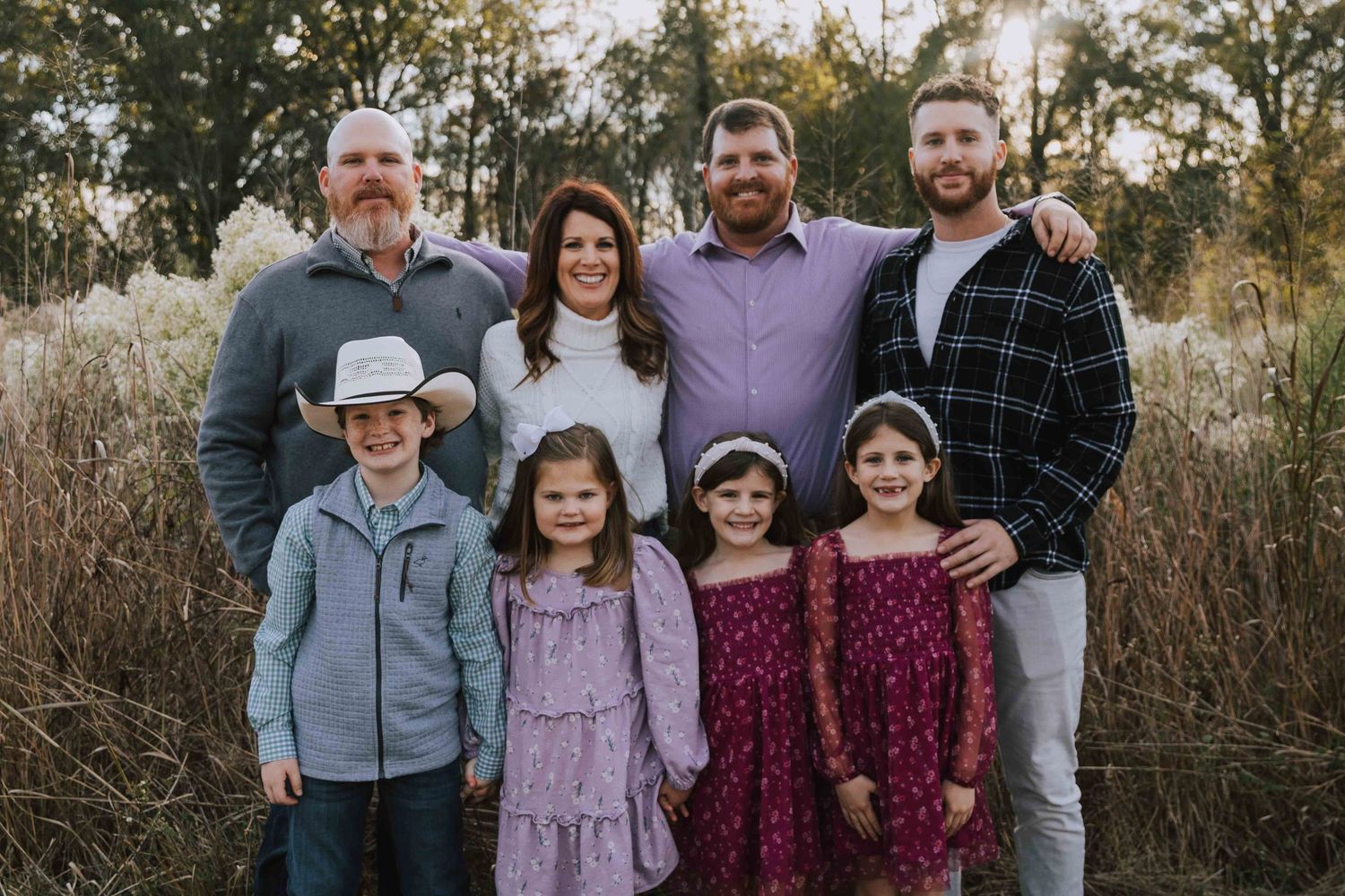 Family of eight posing outdoors in fall setting, adults and children smiling in coordinated outfits.