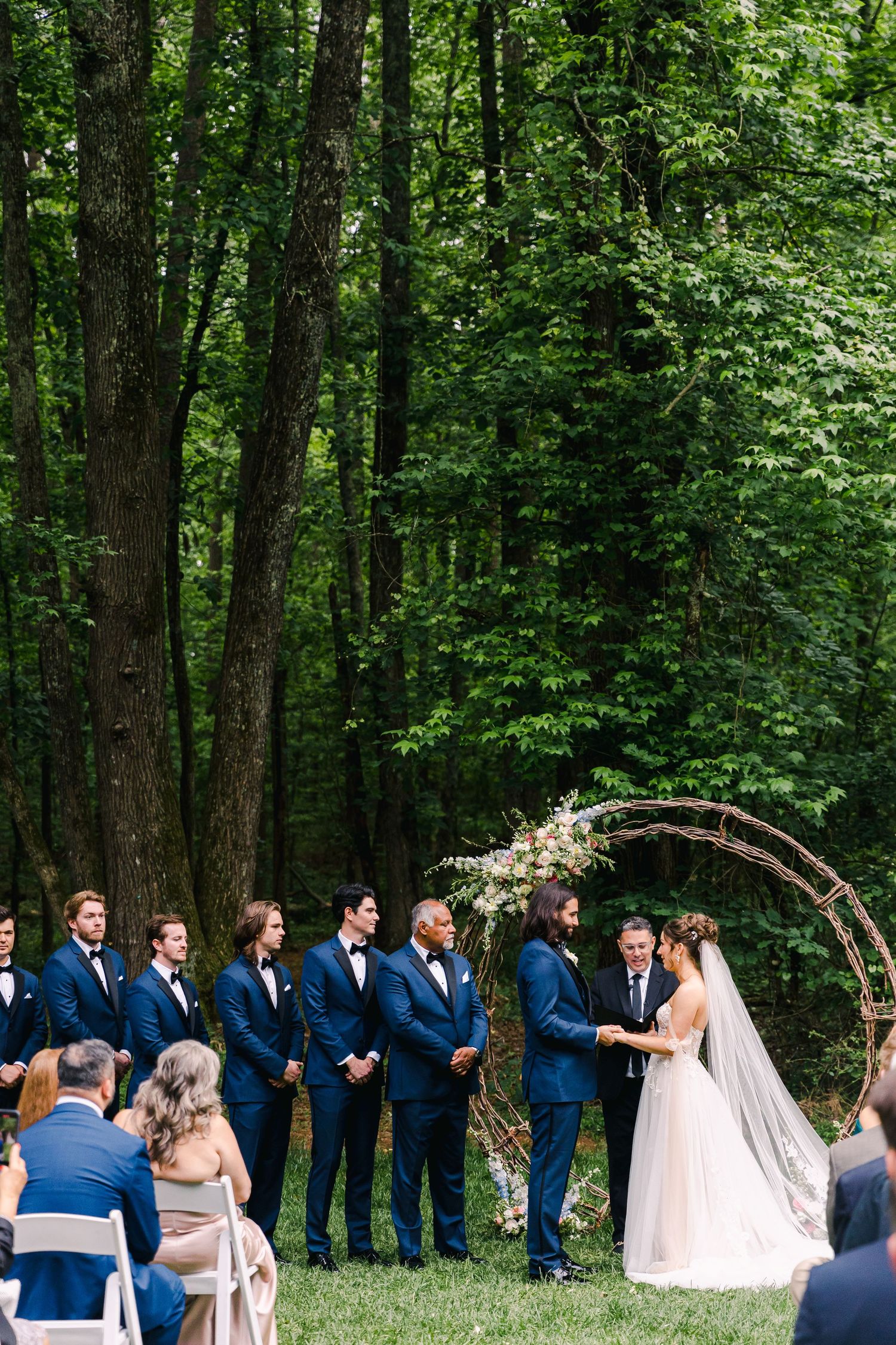 Outdoor wedding ceremony in lush forest with groom, groomsmen in navy suits, bride in white gown, and floral circular arch.