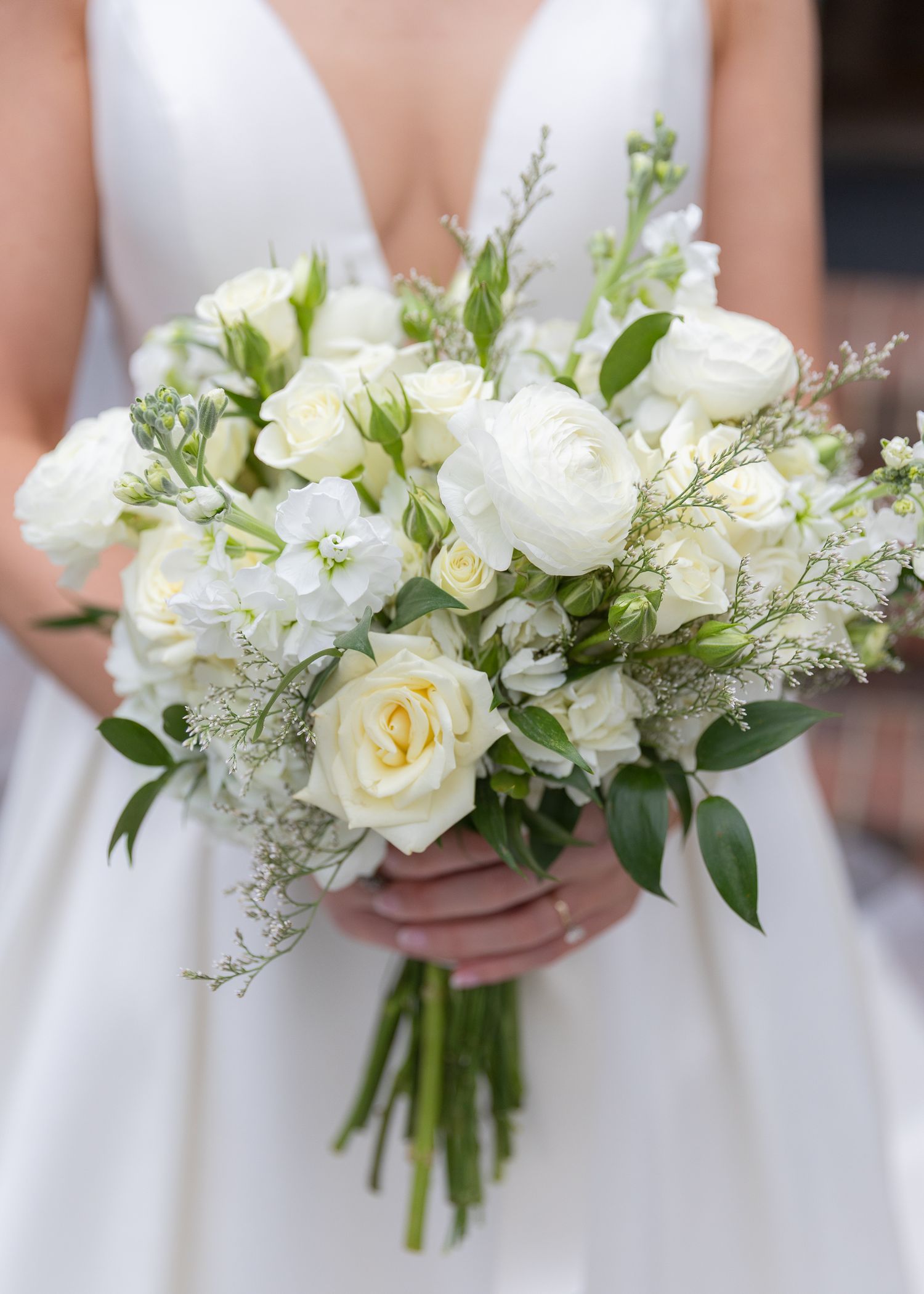Bride holding lush white wedding bouquet with roses, ranunculus, and greenery in ivory dress.