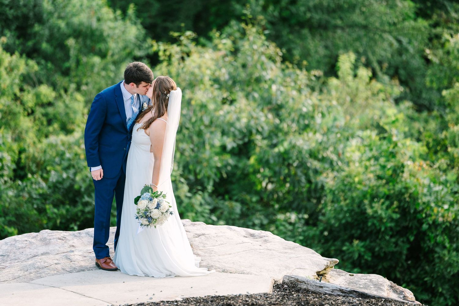 Newlyweds pose on a rocky outcrop surrounded by green summer foliage.