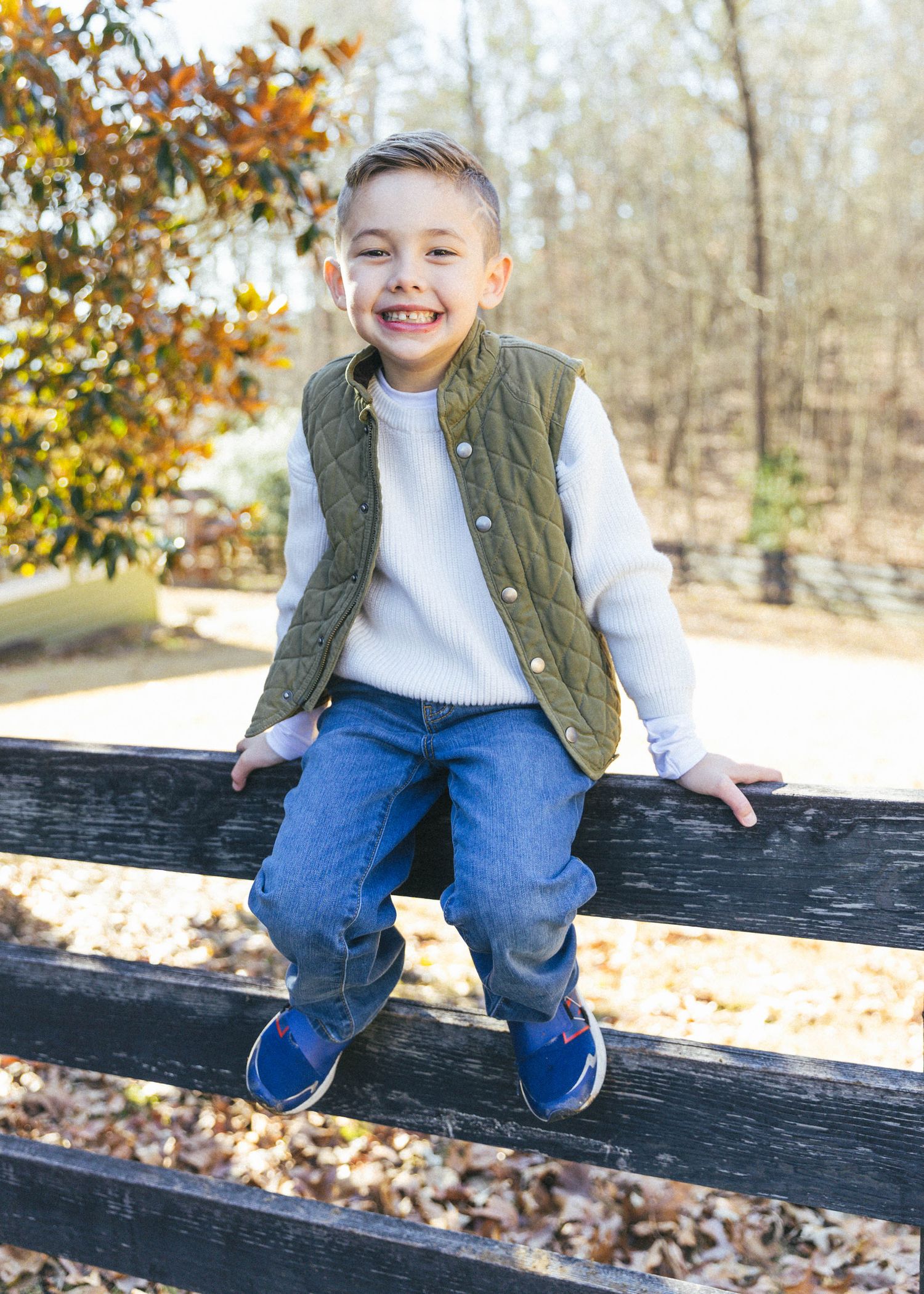 Child in olive vest and jeans sits playfully on wooden fence during autumn.
