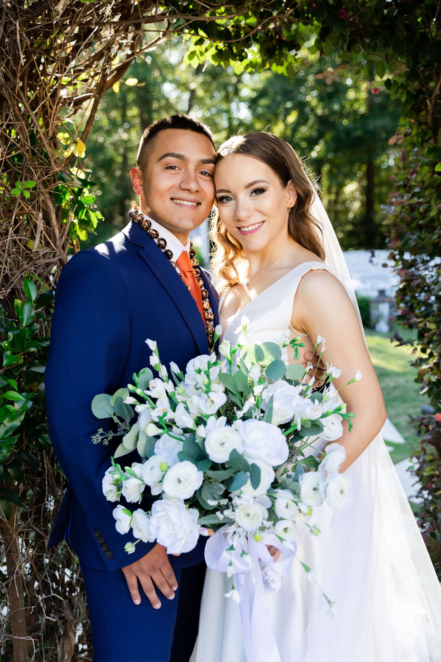 Smiling bride and groom pose outdoors, bride holding white floral bouquet, groom in blue suit with orange tie.