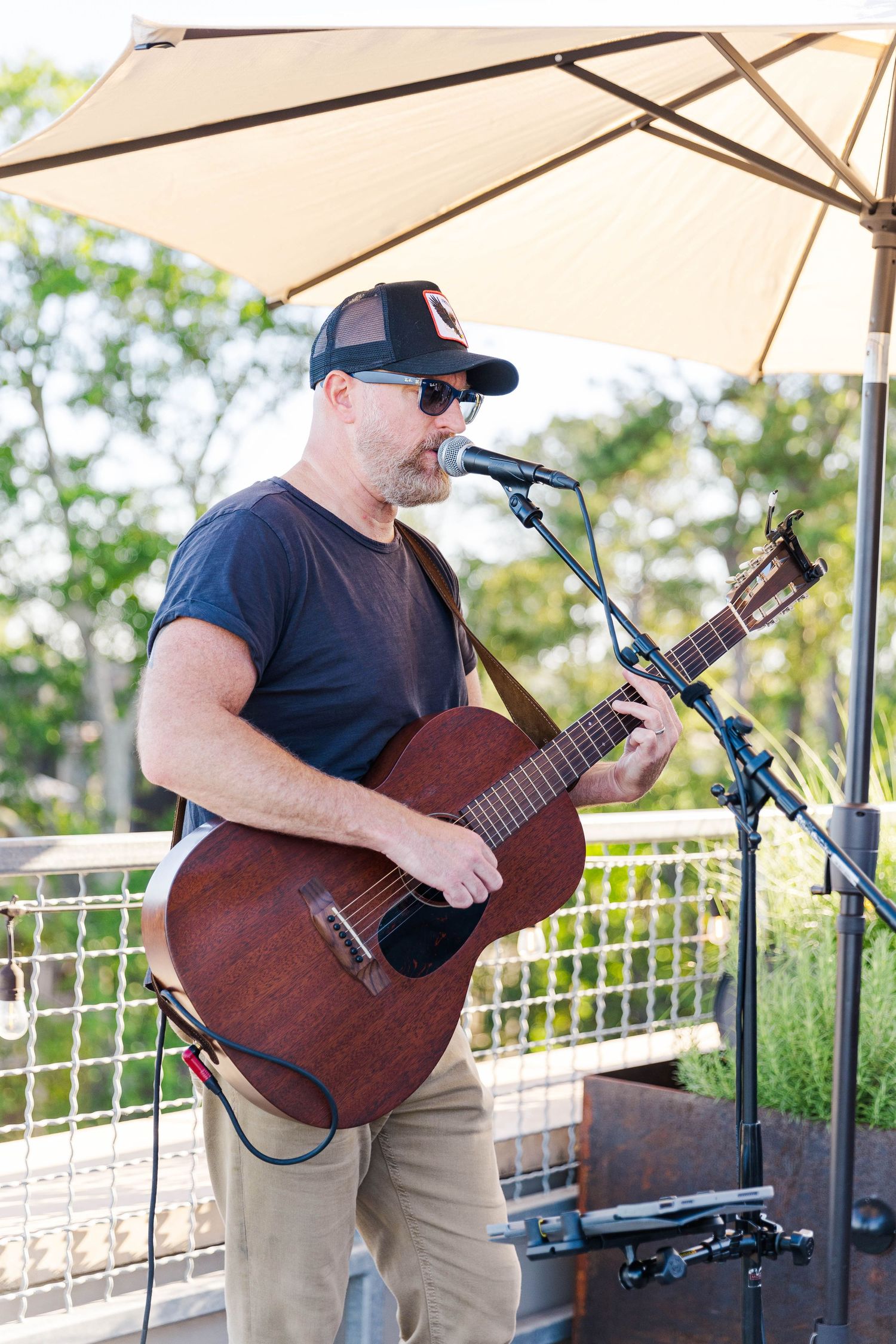 Acoustic guitarist performs on an outdoor stage under a white umbrella during a daytime event.