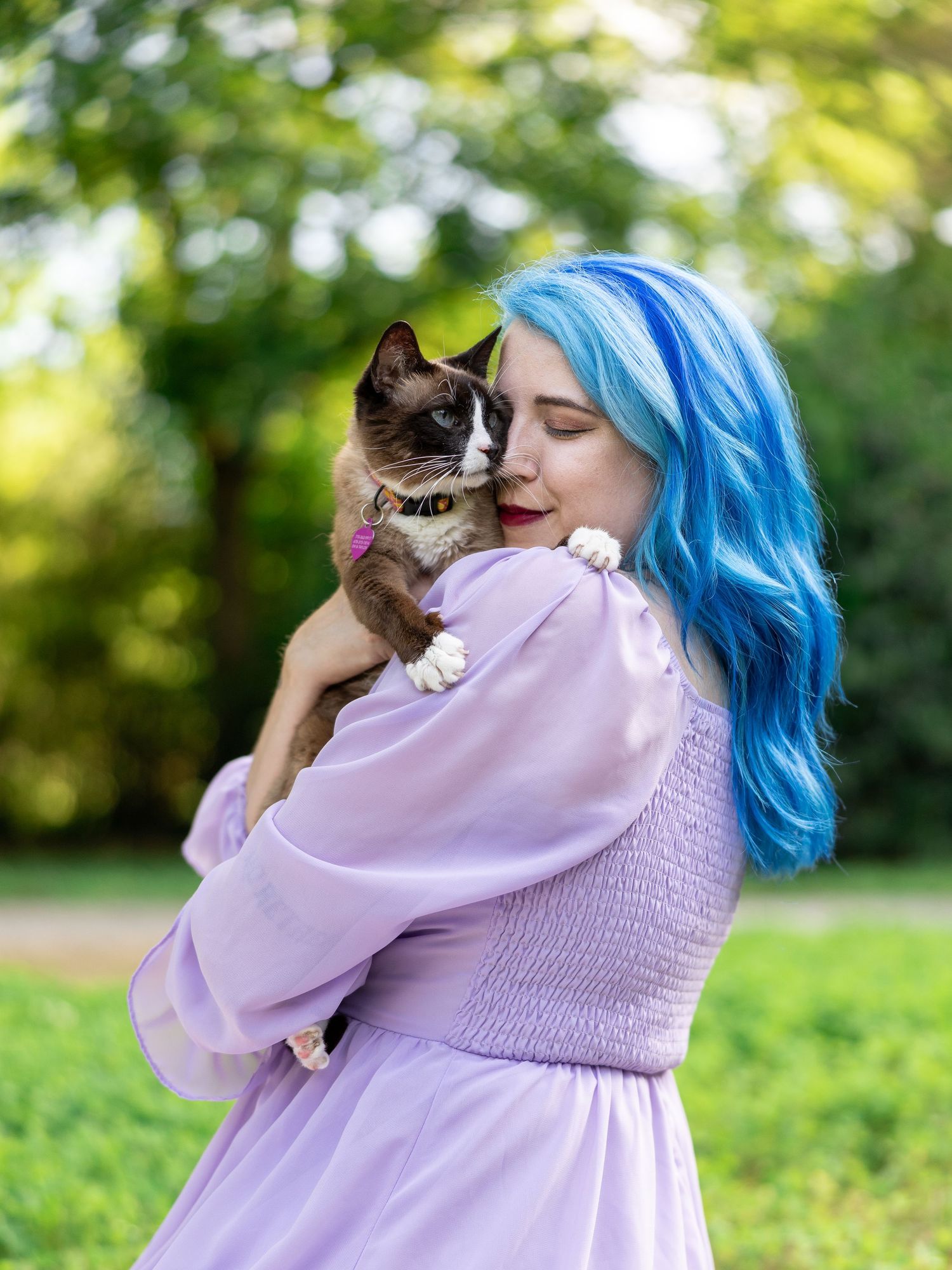 A person with blue hair in a purple dress holding a Siamese cat outdoors.