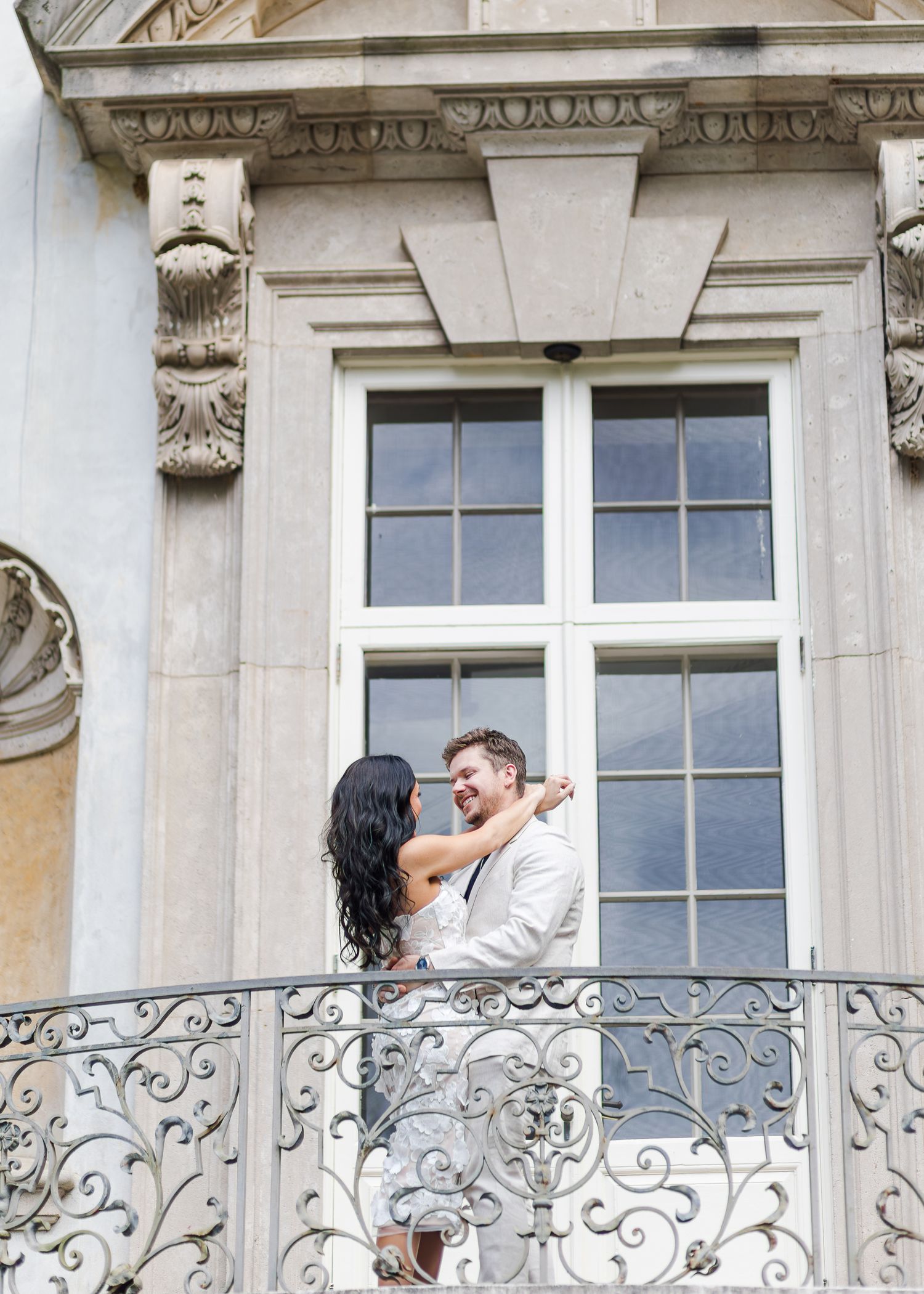 A couple embraces on an ornate wrought iron balcony of an elegant classical building with decorative stonework.