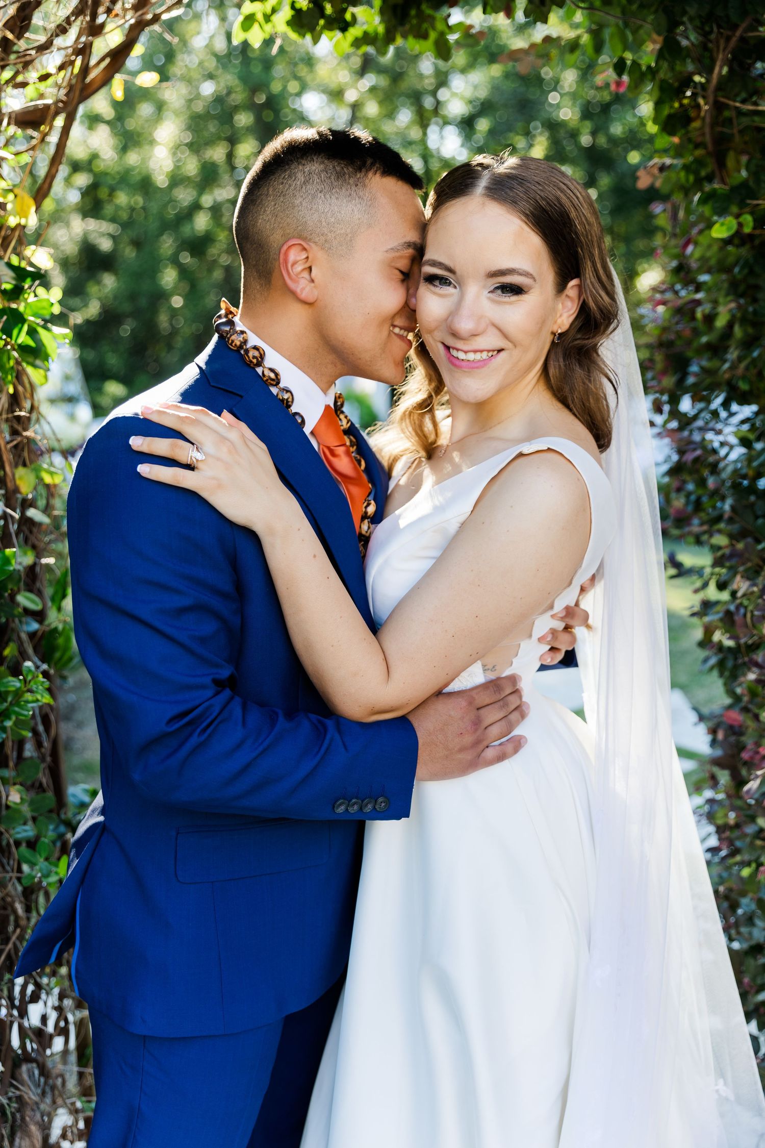 A smiling bride in a white gown and veil embraces her groom in a blue suit outdoors among green foliage.