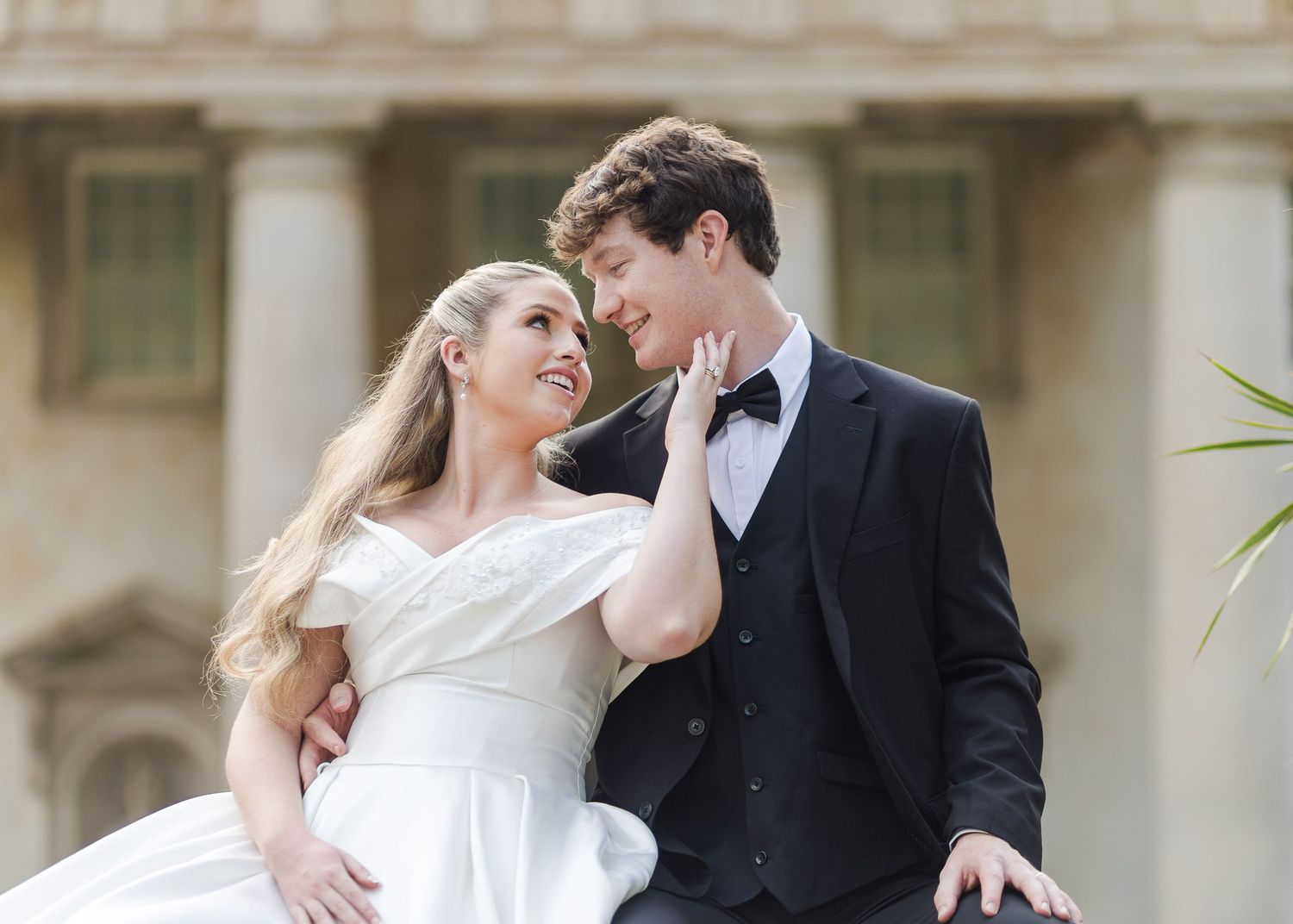 Bride and groom in elegant wedding attire share a romantic moment outdoors near classical stone columns.
