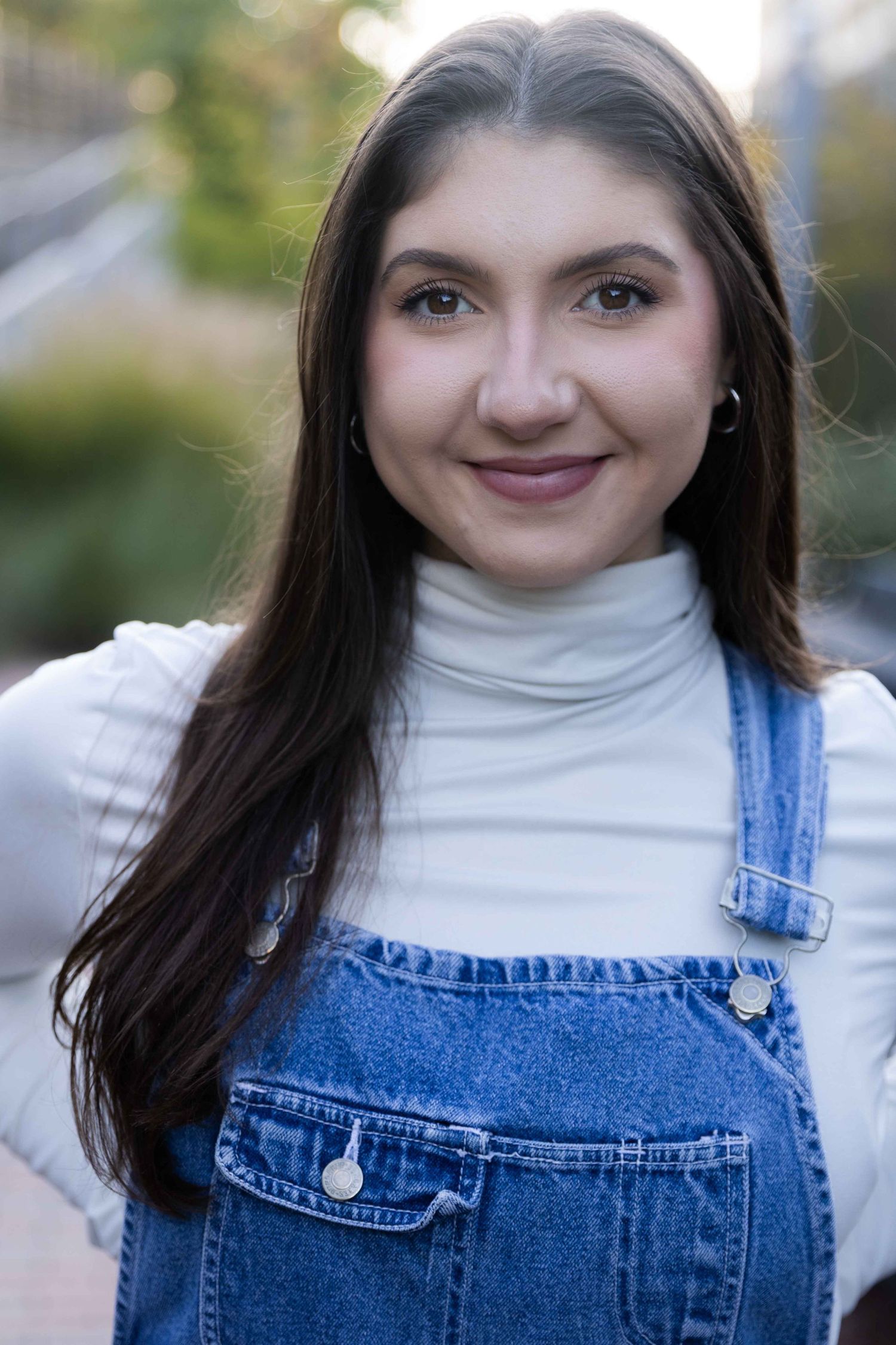 Young woman with long dark hair smiling, wearing denim overalls over a white turtleneck outdoors.