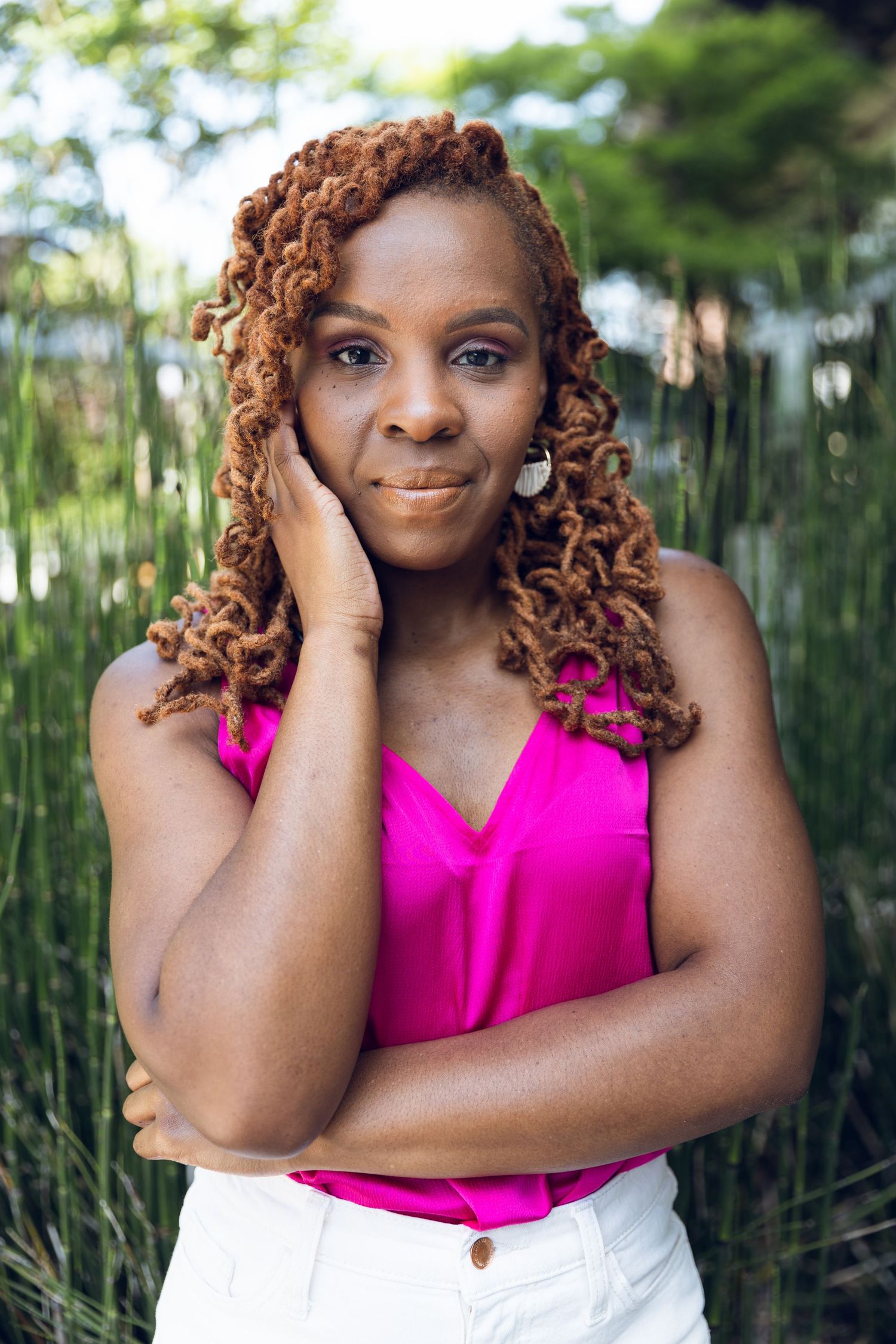 A Black woman with auburn locs wearing a pink top and white jeans poses outdoors against lush green foliage.
