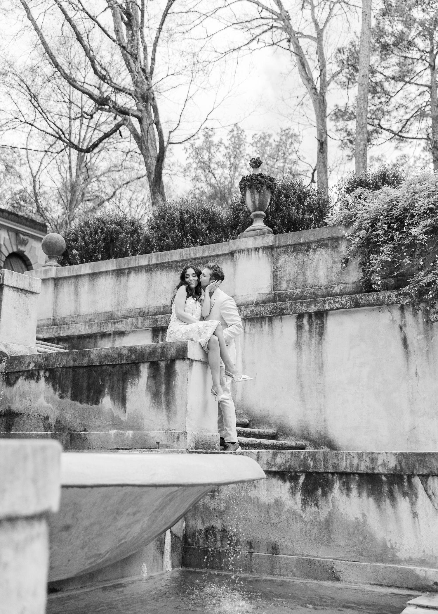 Black and white photo of a couple sitting on a stone fountain wall surrounded by trees and lush hedges at the Swan House in Atlanta, Georgia