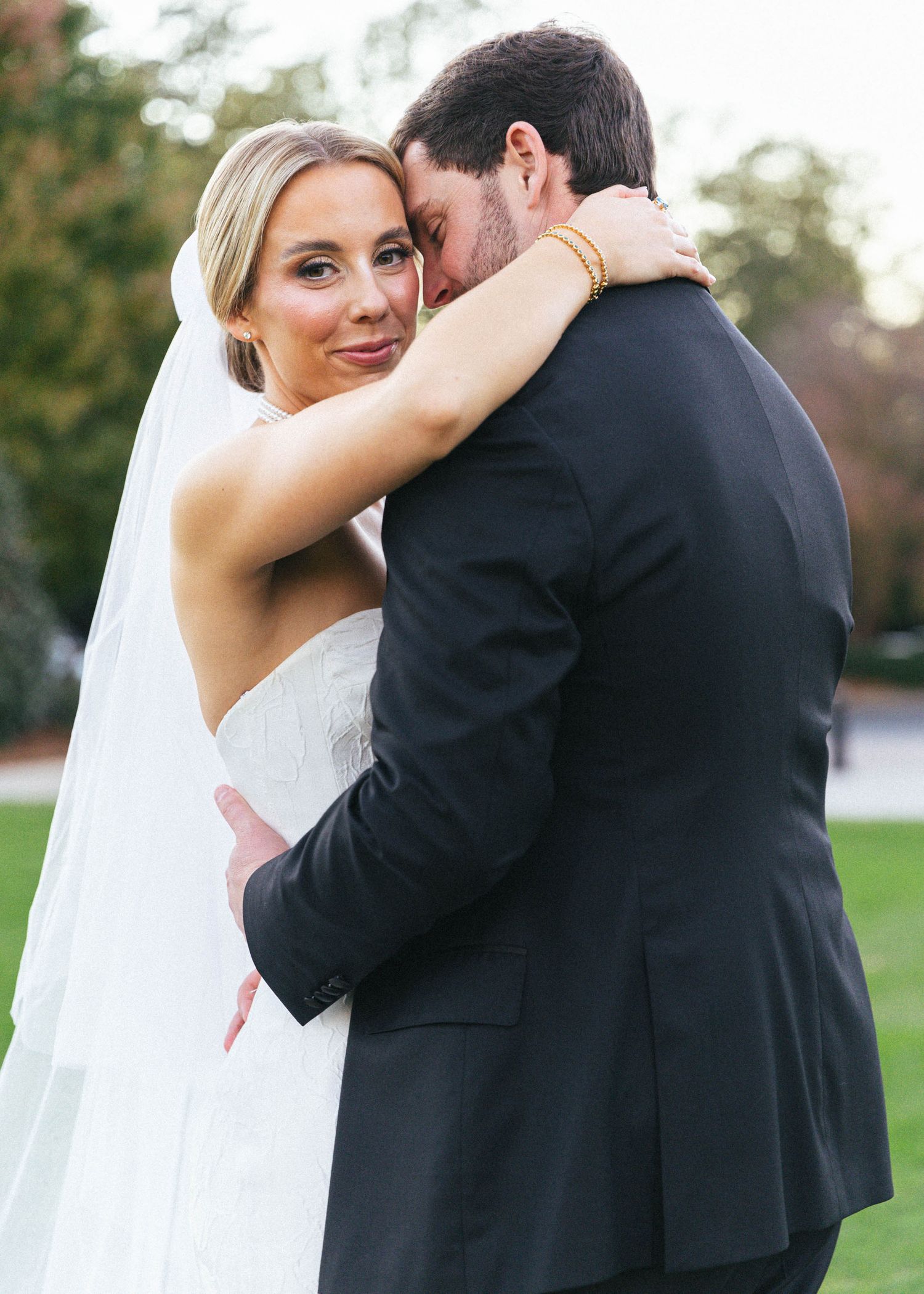 Bride in white gown and veil embraces groom in black suit outdoors at a wedding ceremony.
