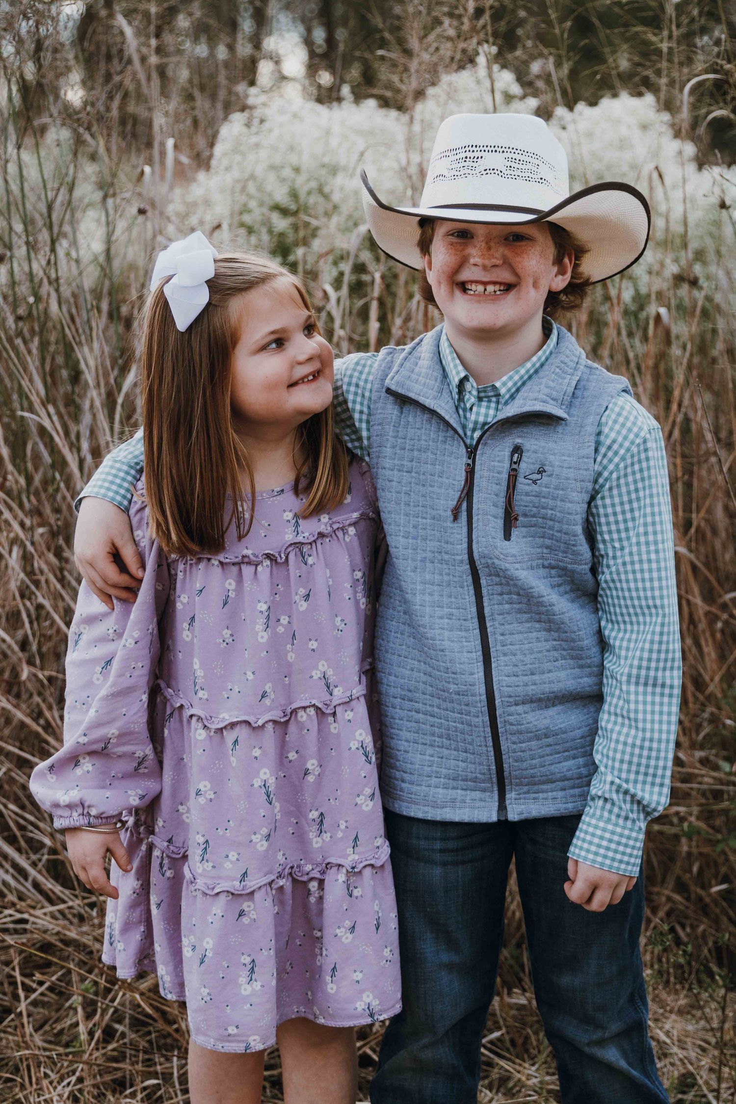 Two smiling children pose outdoors, a girl in a purple dress with a bow and a boy in a cowboy hat and blue vest.
