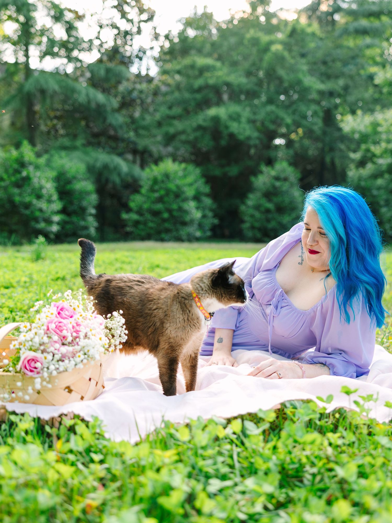 Person with blue hair relaxes on picnic blanket with cat in sunny garden setting.
