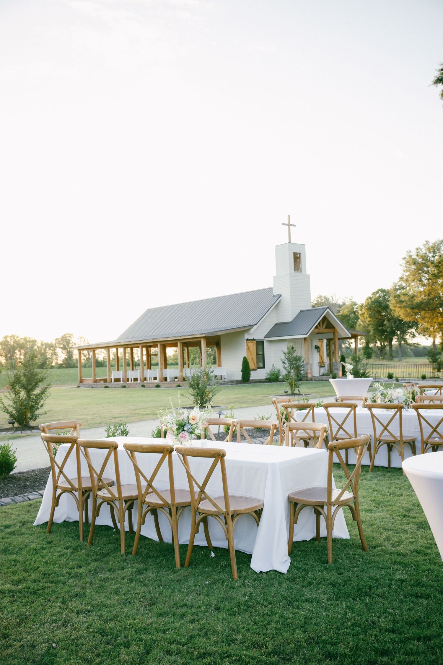 Outdoor wedding reception with wooden cross-back chairs and white linen tables on green lawn beside a charming white chapel.