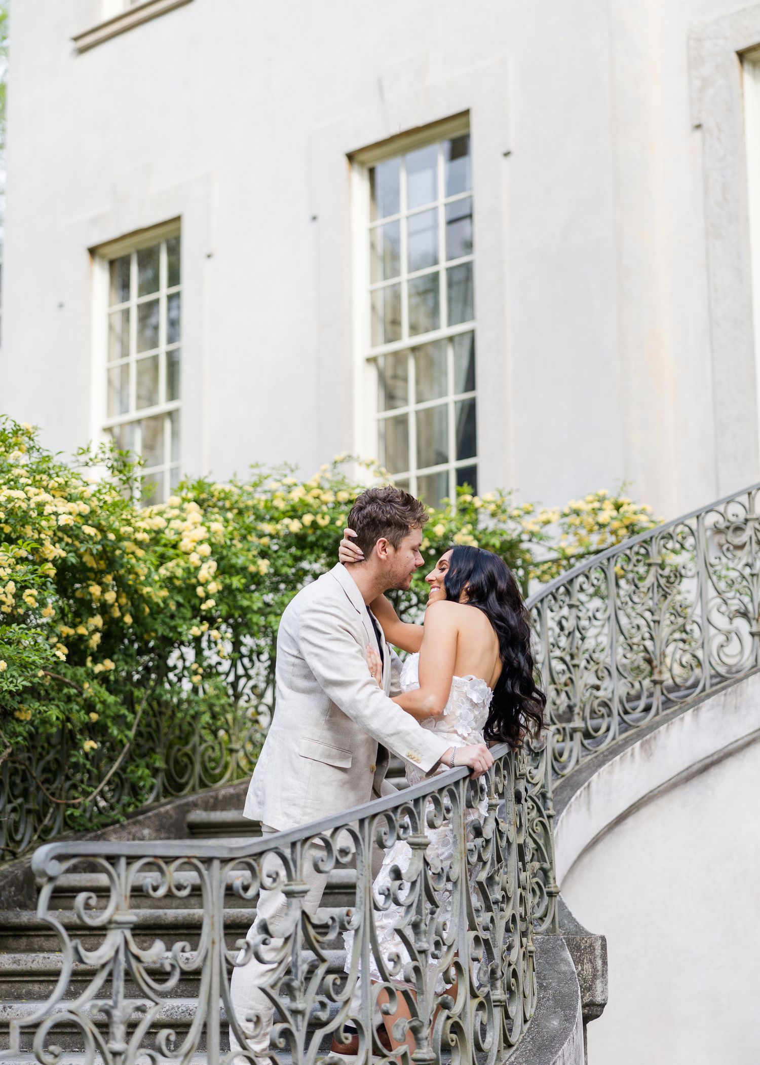 Couple sharing a romantic kiss on ornate iron staircase outside elegant white mansion with yellow roses.
