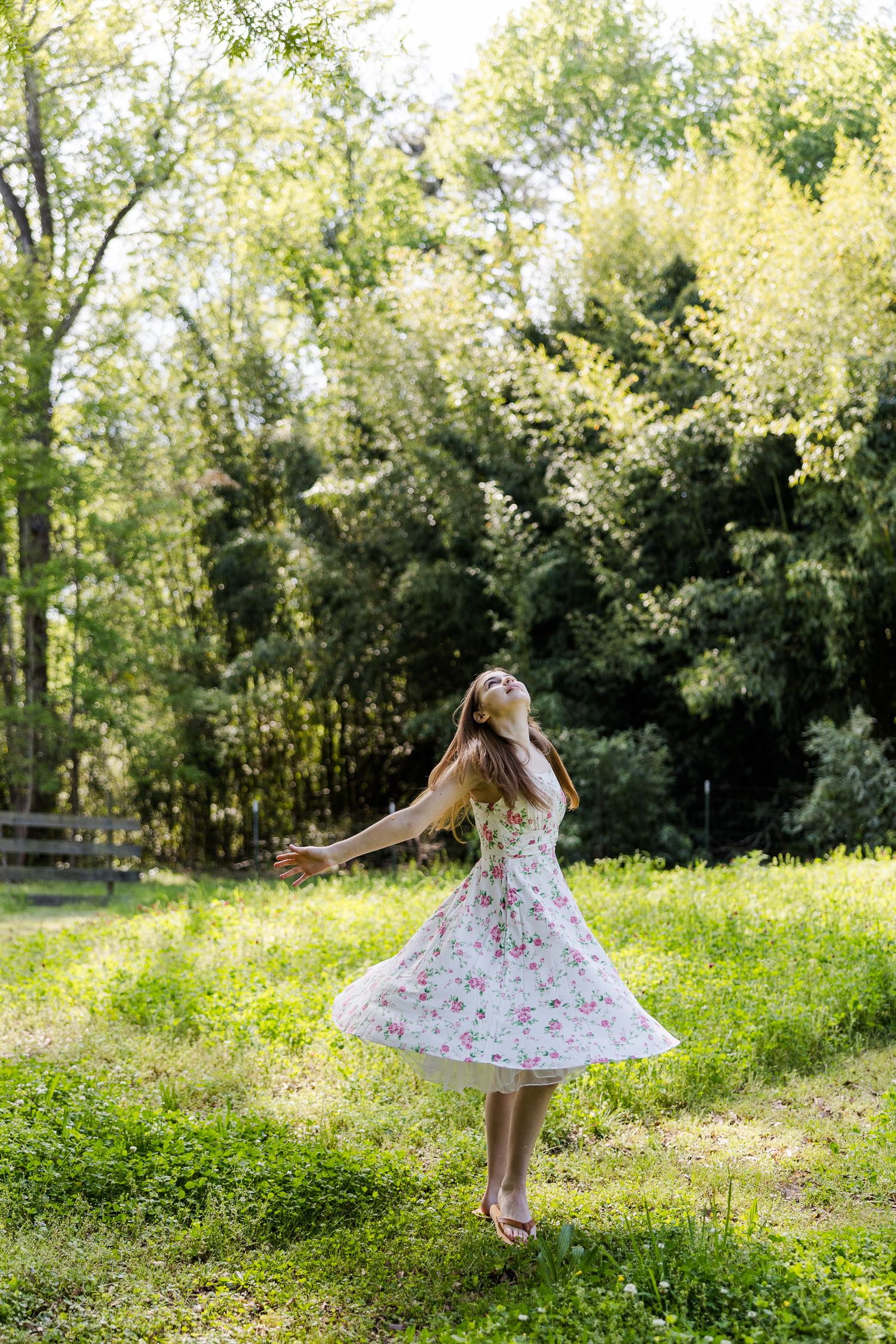 Young woman in floral dress twirling joyfully in lush green garden surrounded by trees on a sunny day.