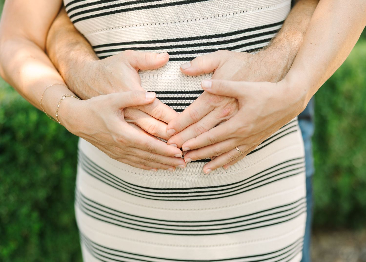 Couple forming a heart shape with hands on pregnant belly wearing striped dress outdoors.