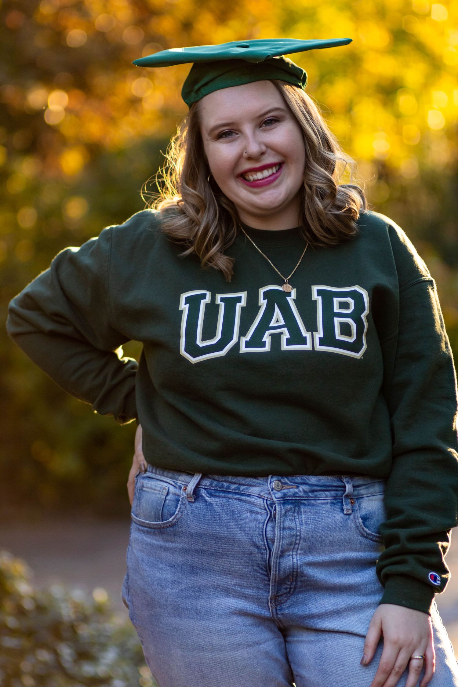 A person wearing a dark green university sweatshirt and jeans poses outdoors against a golden autumn background.