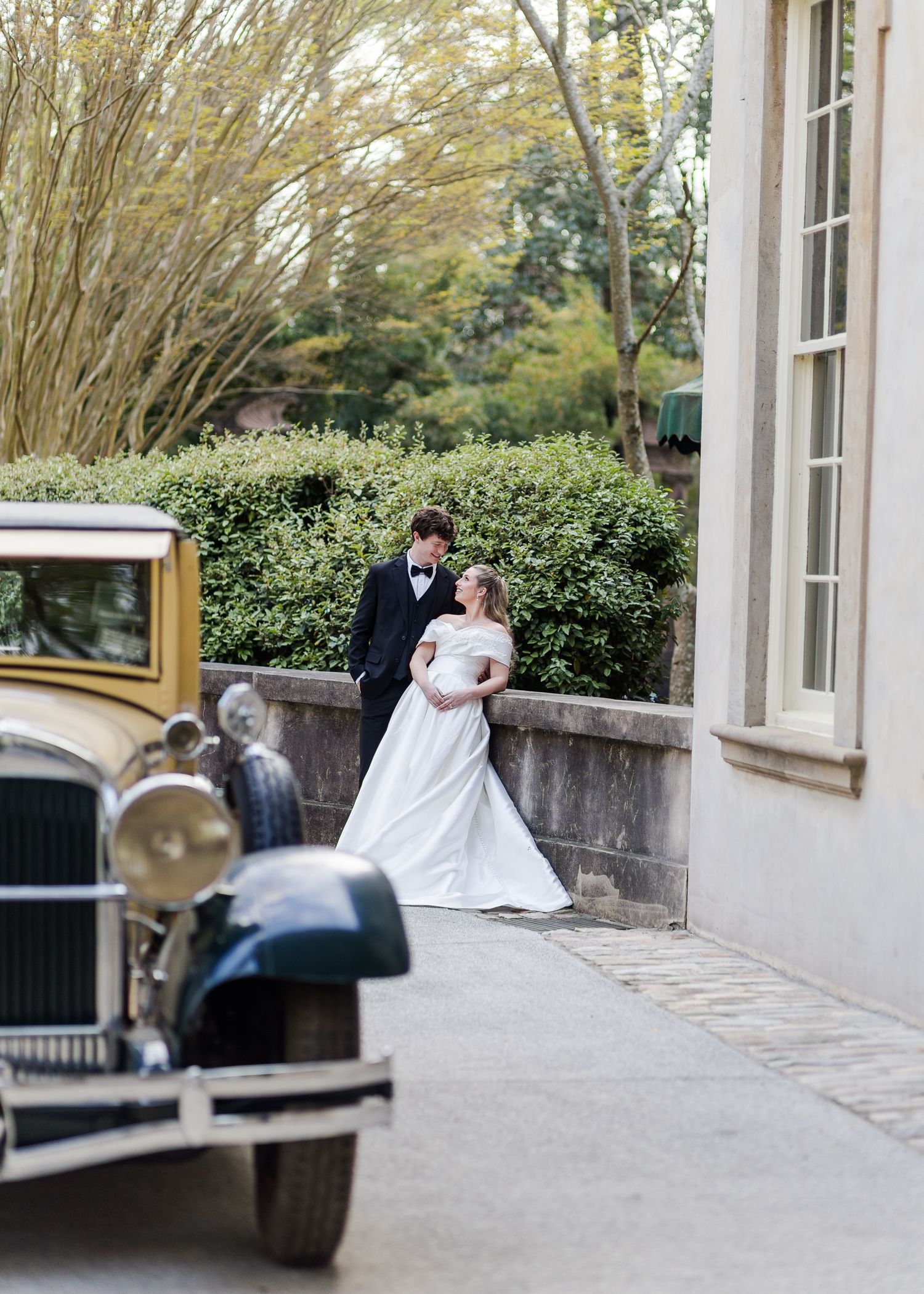 Bride and groom embrace near a vintage classic car outside a historic stone wedding venue.