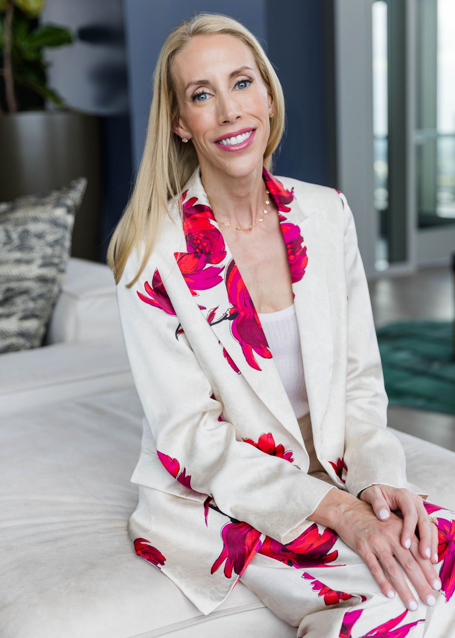 Woman in white floral blazer poses gracefully on light colored furniture.