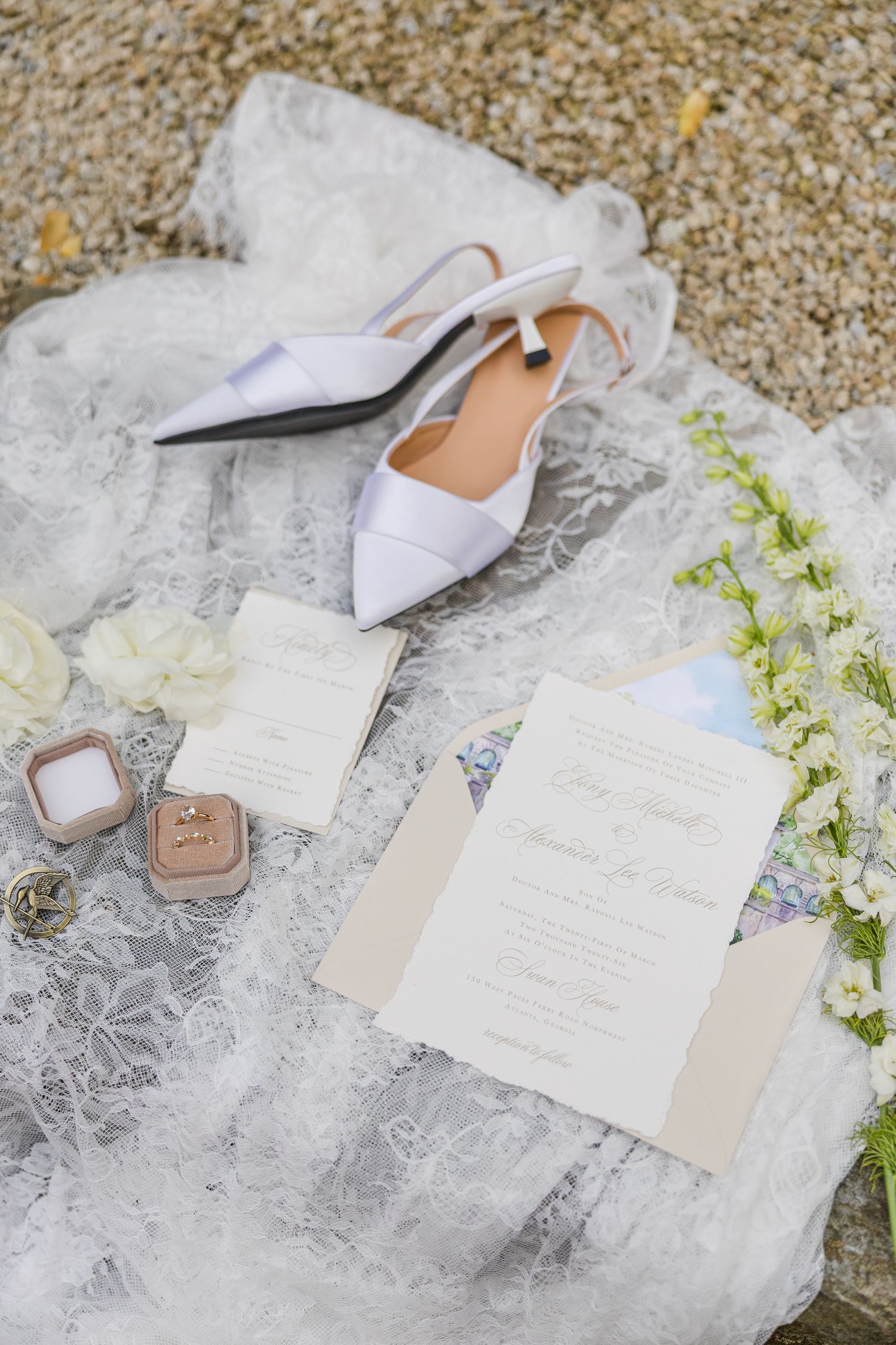 White wedding heels, ring box, floral invitations, and lace veil arranged as a bridal flat lay on stone.