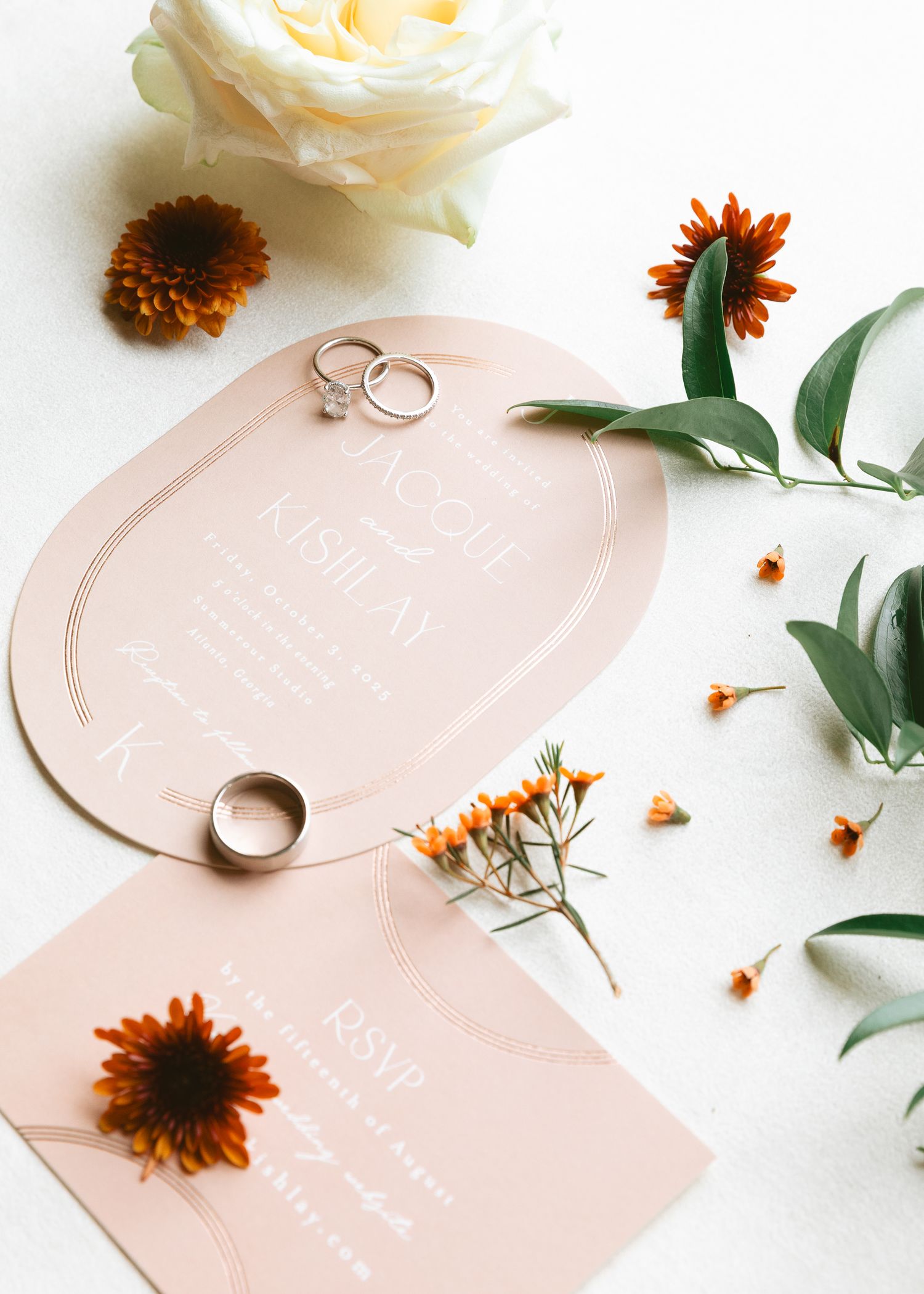 Elegant wedding invitation suite with gold rings, blush pink cards, white roses, and rustic orange flowers on white background.
