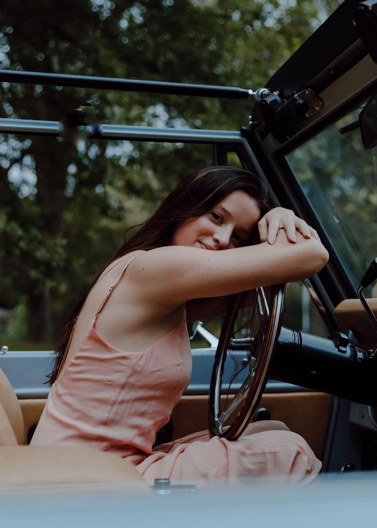 Young woman in pink dress leaning on steering wheel of vintage open-top vehicle, smiling outdoors.