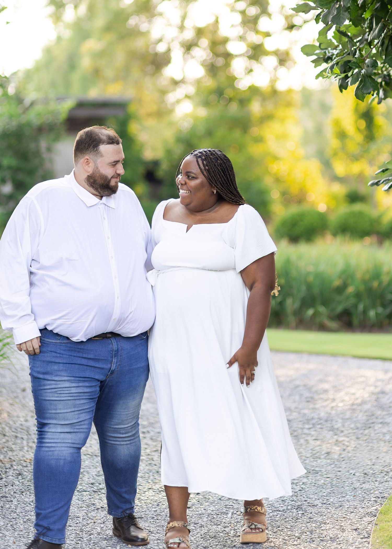 A couple in matching white outfits walk together on a gravel path surrounded by lush greenery.