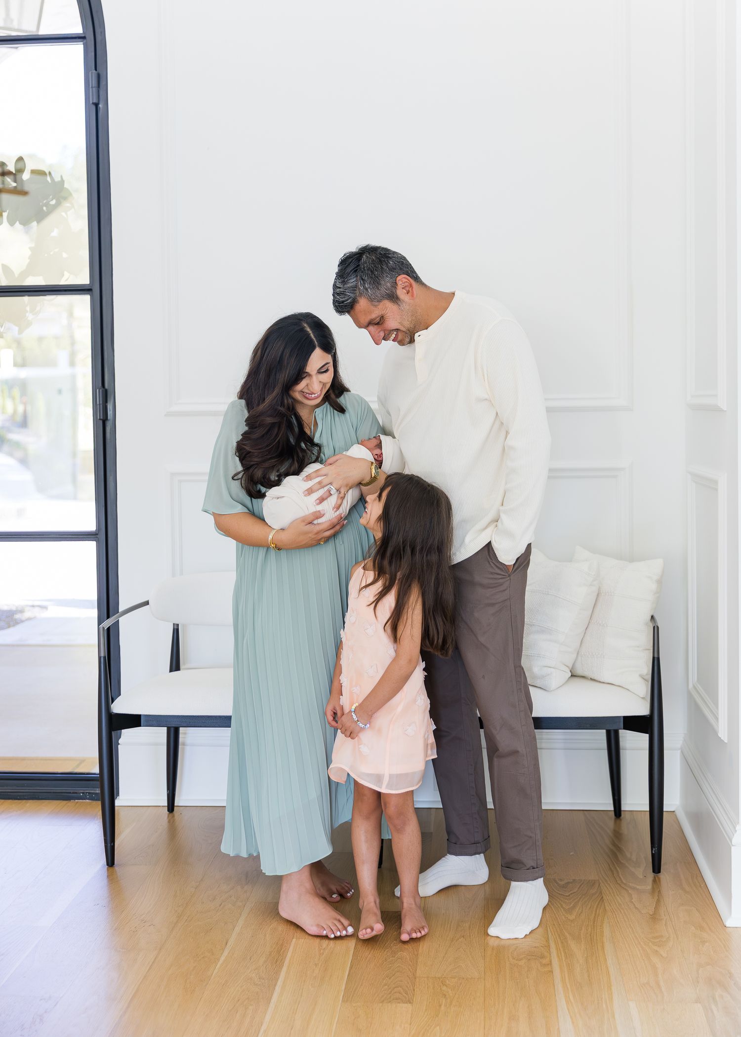 A family of four gazes lovingly at their newborn baby in a bright, elegant white room with hardwood floors.