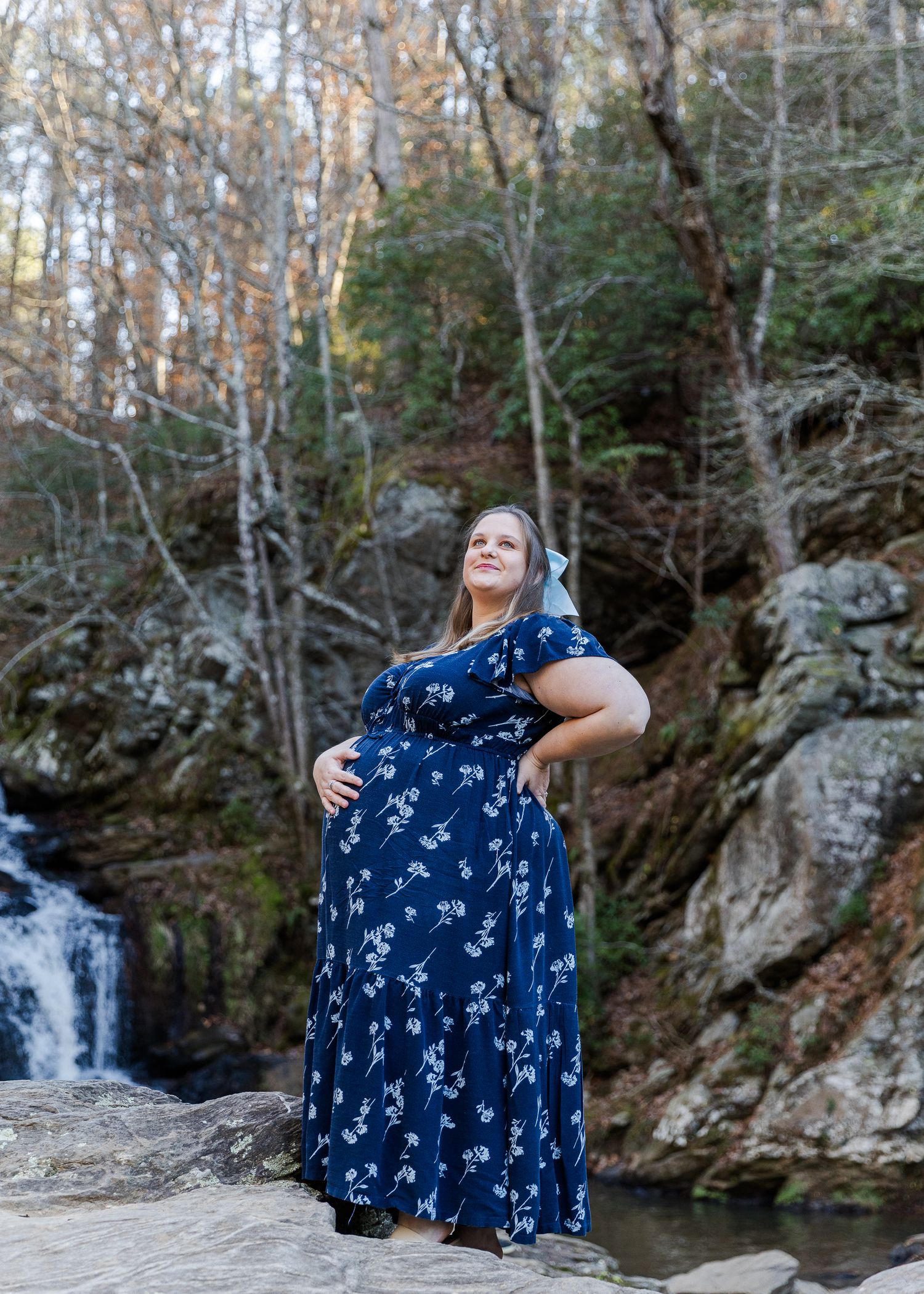 Pregnant woman in navy floral maxi dress poses on rocks near waterfall surrounded by autumn trees.