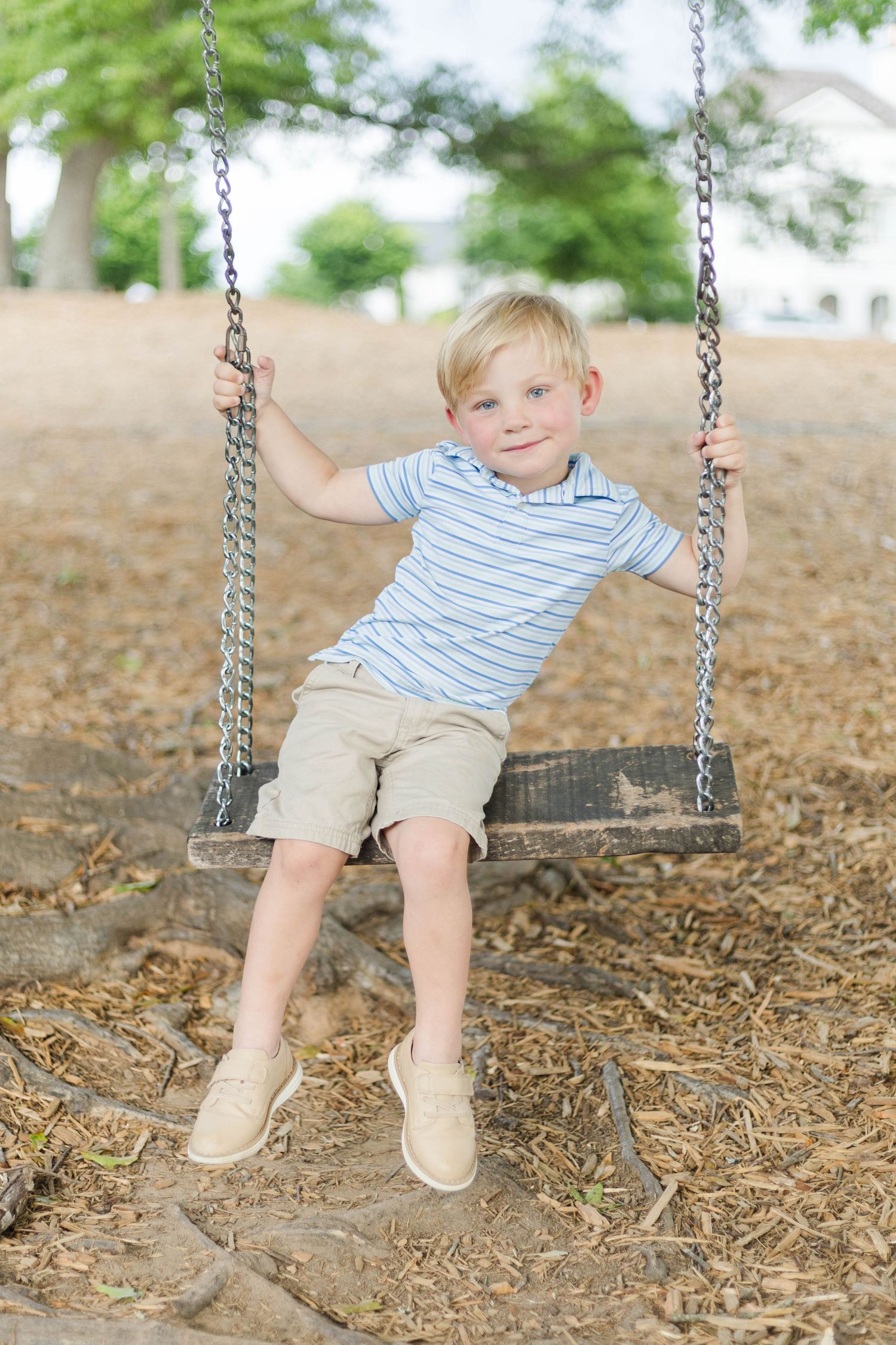 Young blonde boy sitting on a wooden chain swing at a playground, smiling at the camera on a sunny day.