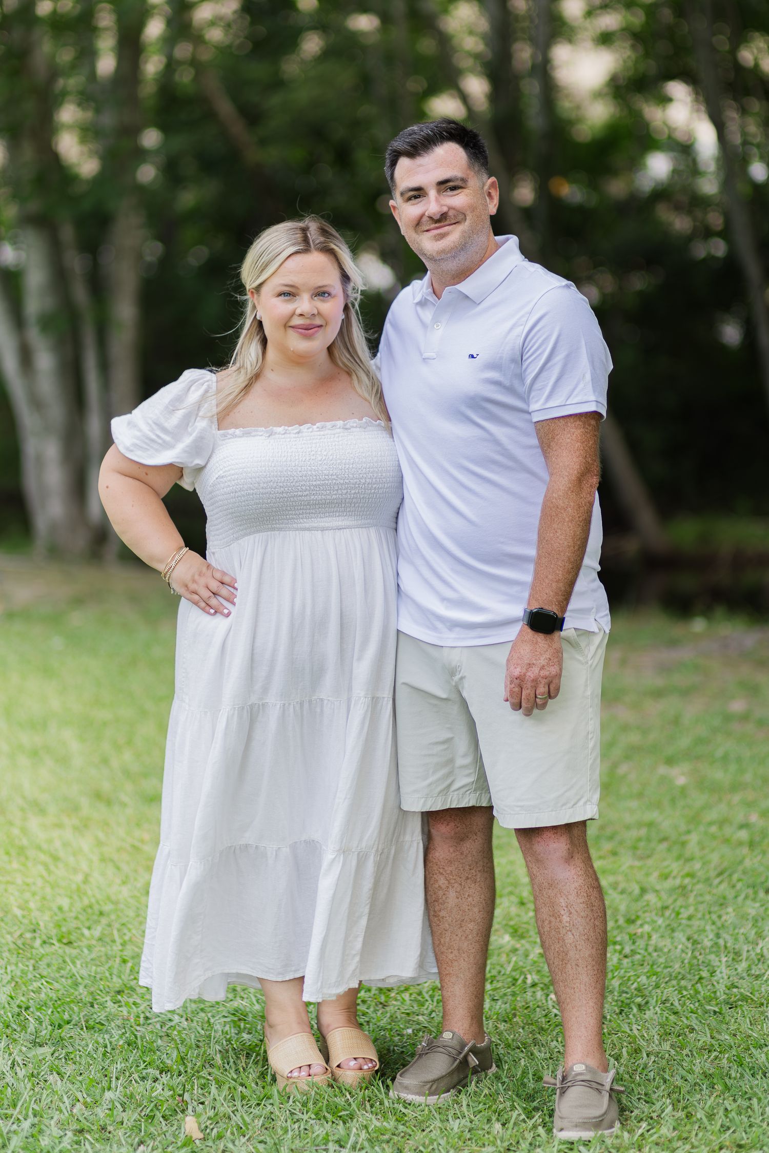 Couple posing outdoors on green grass wearing white outfits, woman in dress and man in polo shirt and shorts.