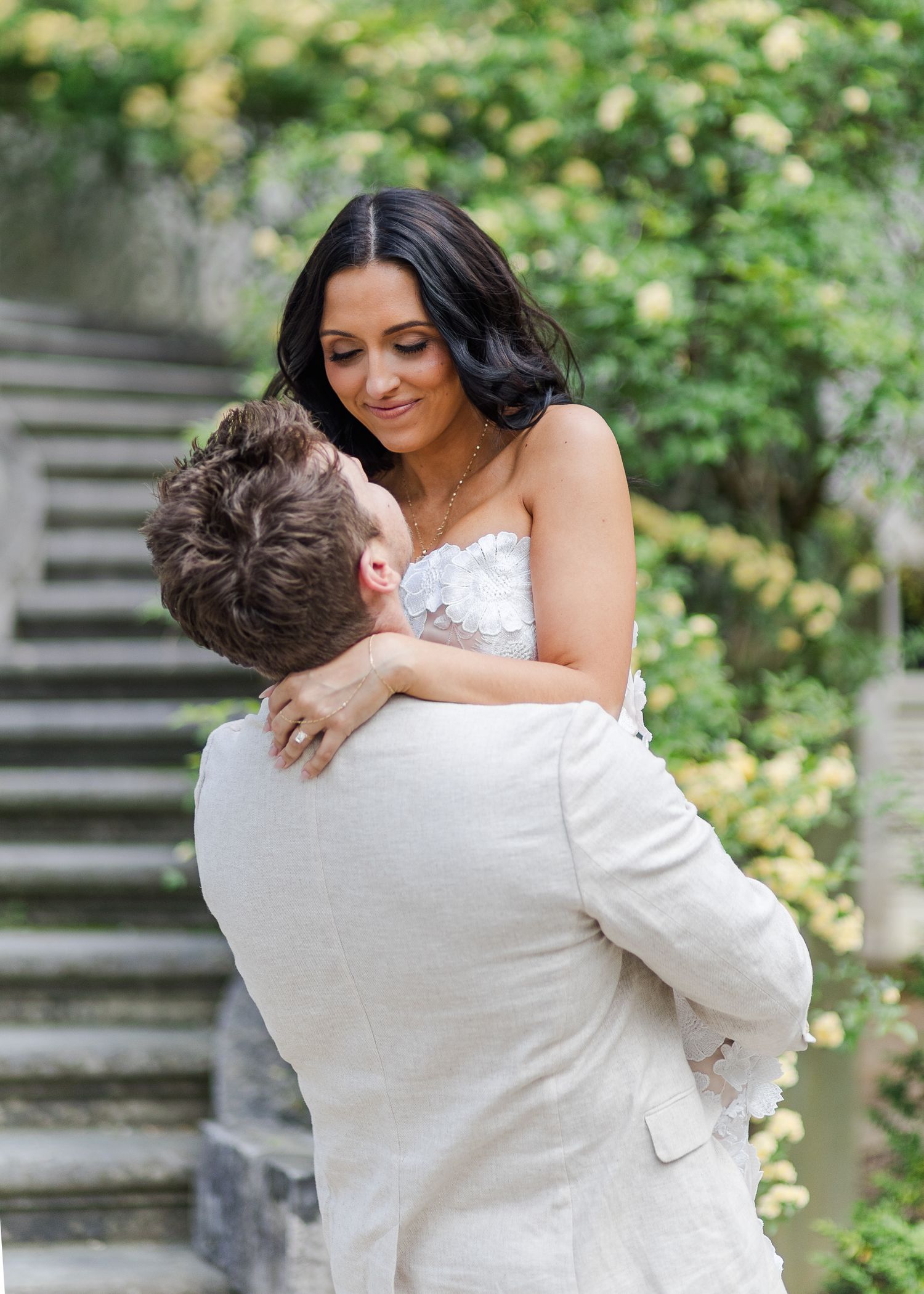 Groom lifting bride in white lace dress outdoors, surrounded by lush greenery and stone steps.