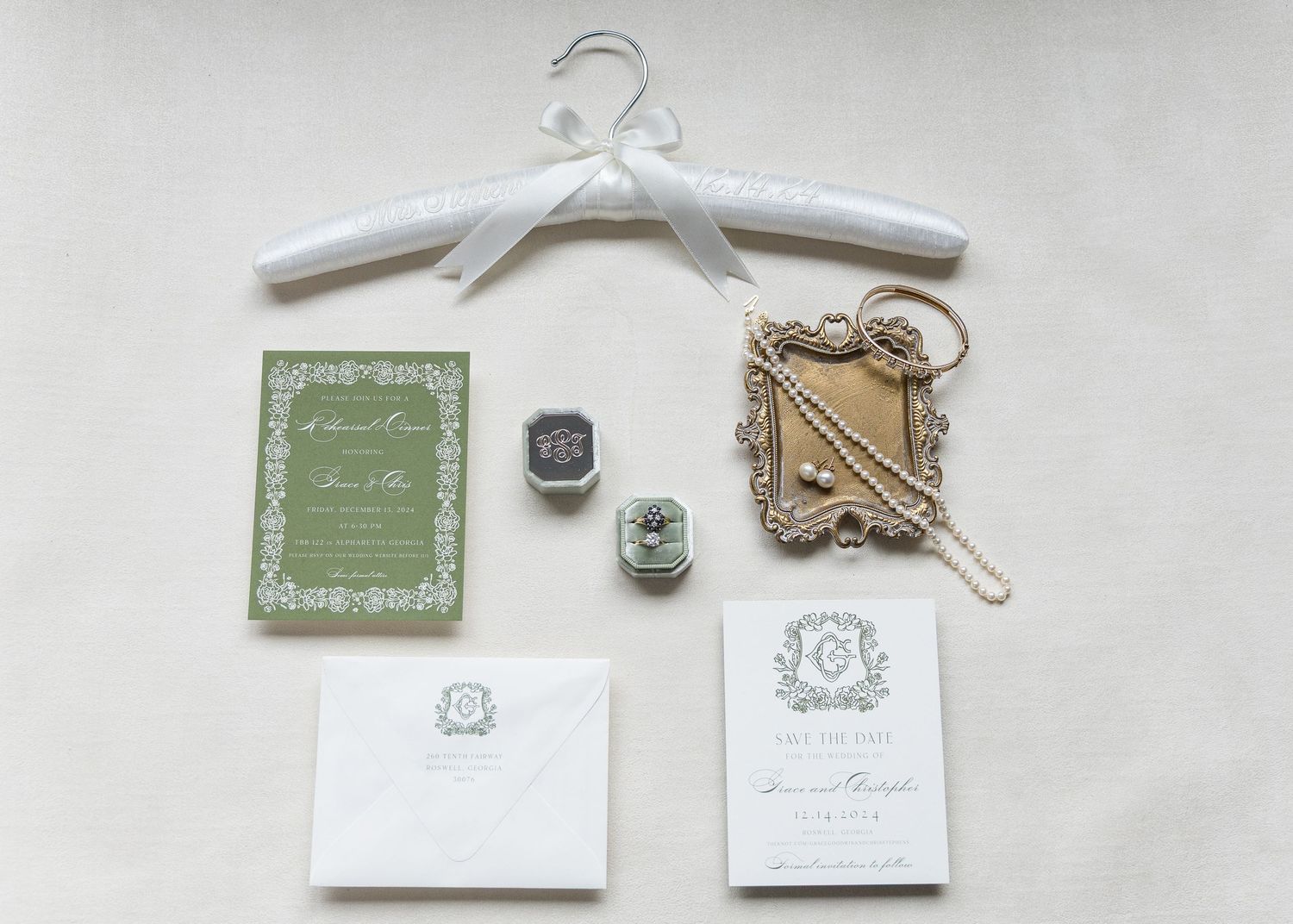 Elegant wedding stationery flat lay with green invite, wax seal, ornate mirror, and monogram envelope on neutral background.