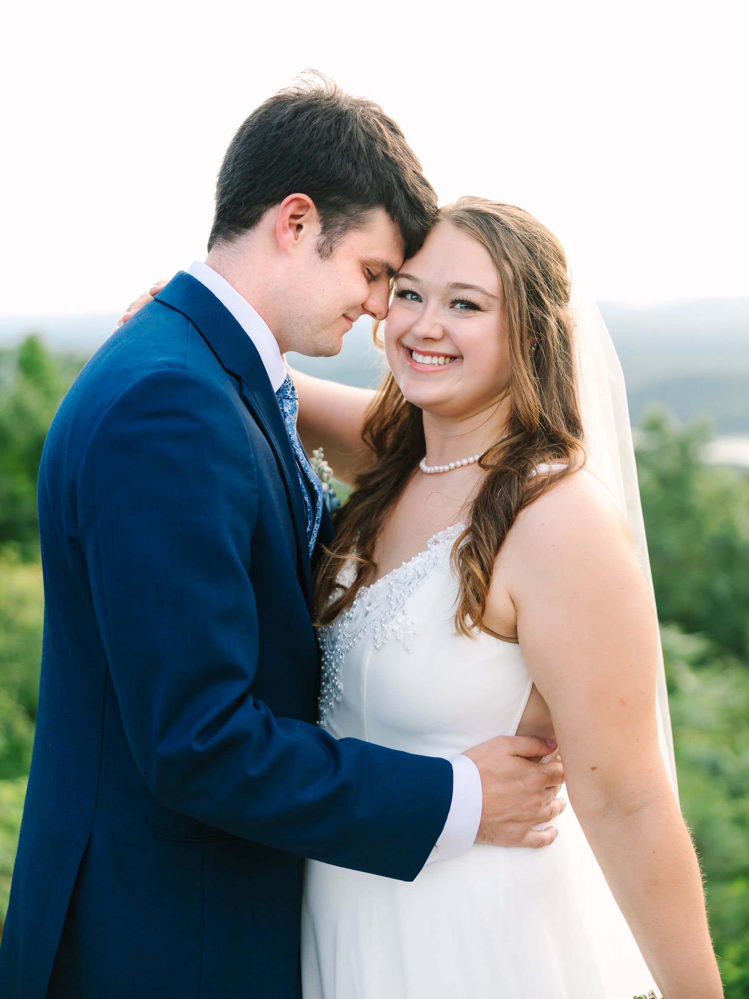A bride in a white gown and veil smiles as she embraces a groom in a navy suit outdoors at a wedding.
