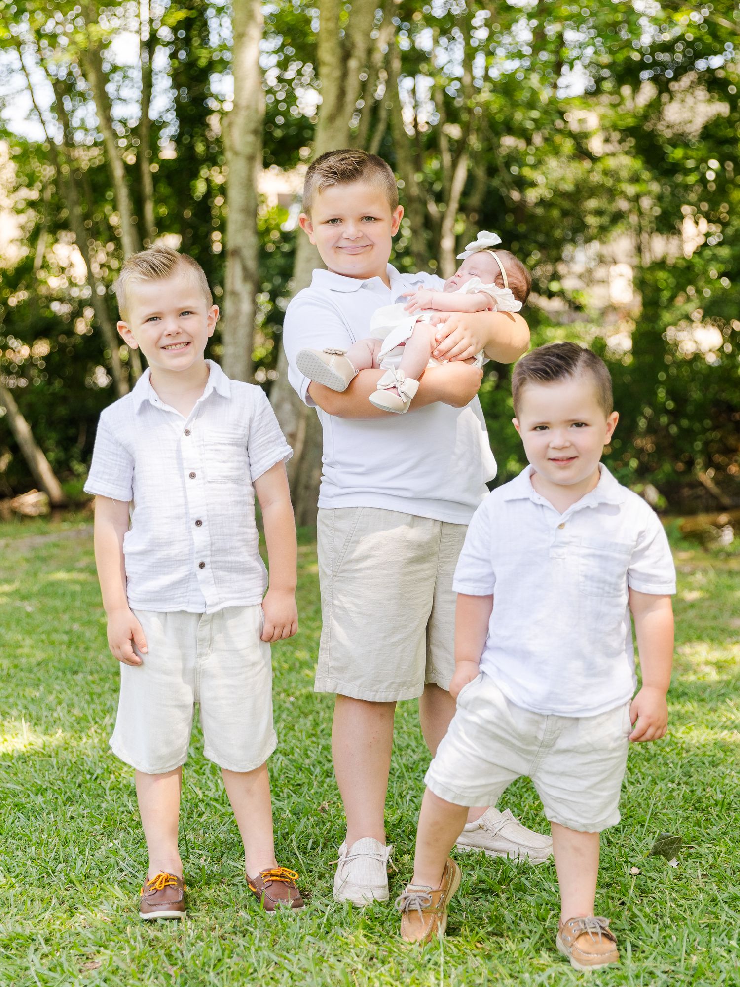 Three young boys in white shirts and khaki shorts pose outdoors, with the oldest holding a newborn baby.