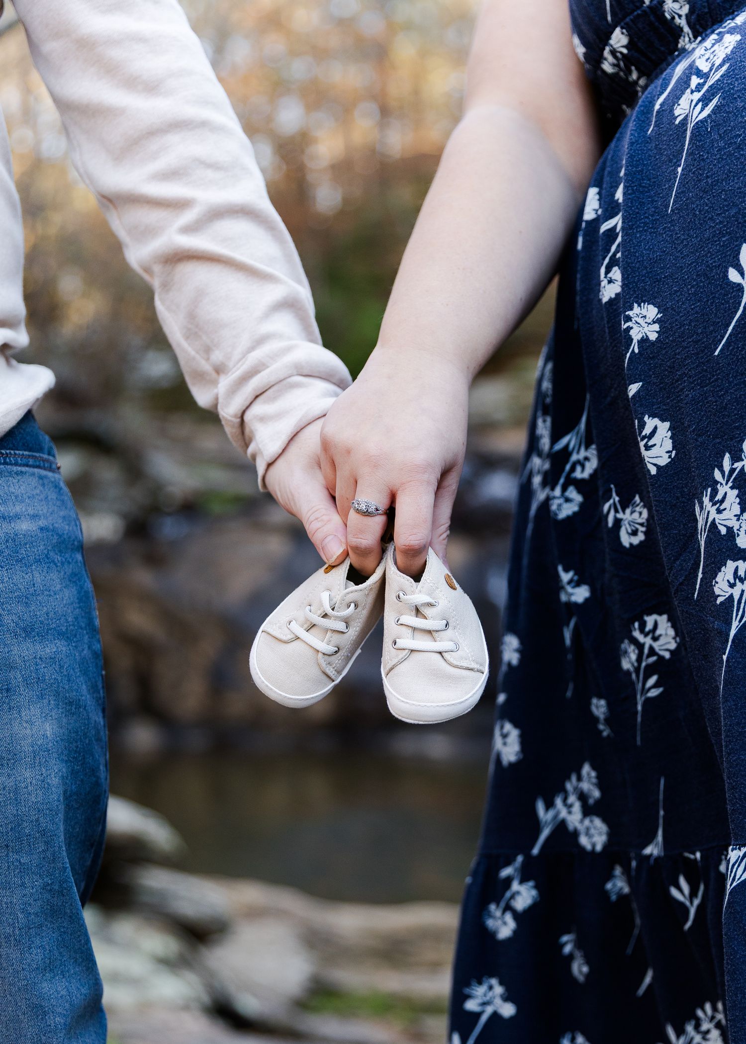 Couple holding tiny white baby sneakers together outdoors, announcing pregnancy near a rocky stream.