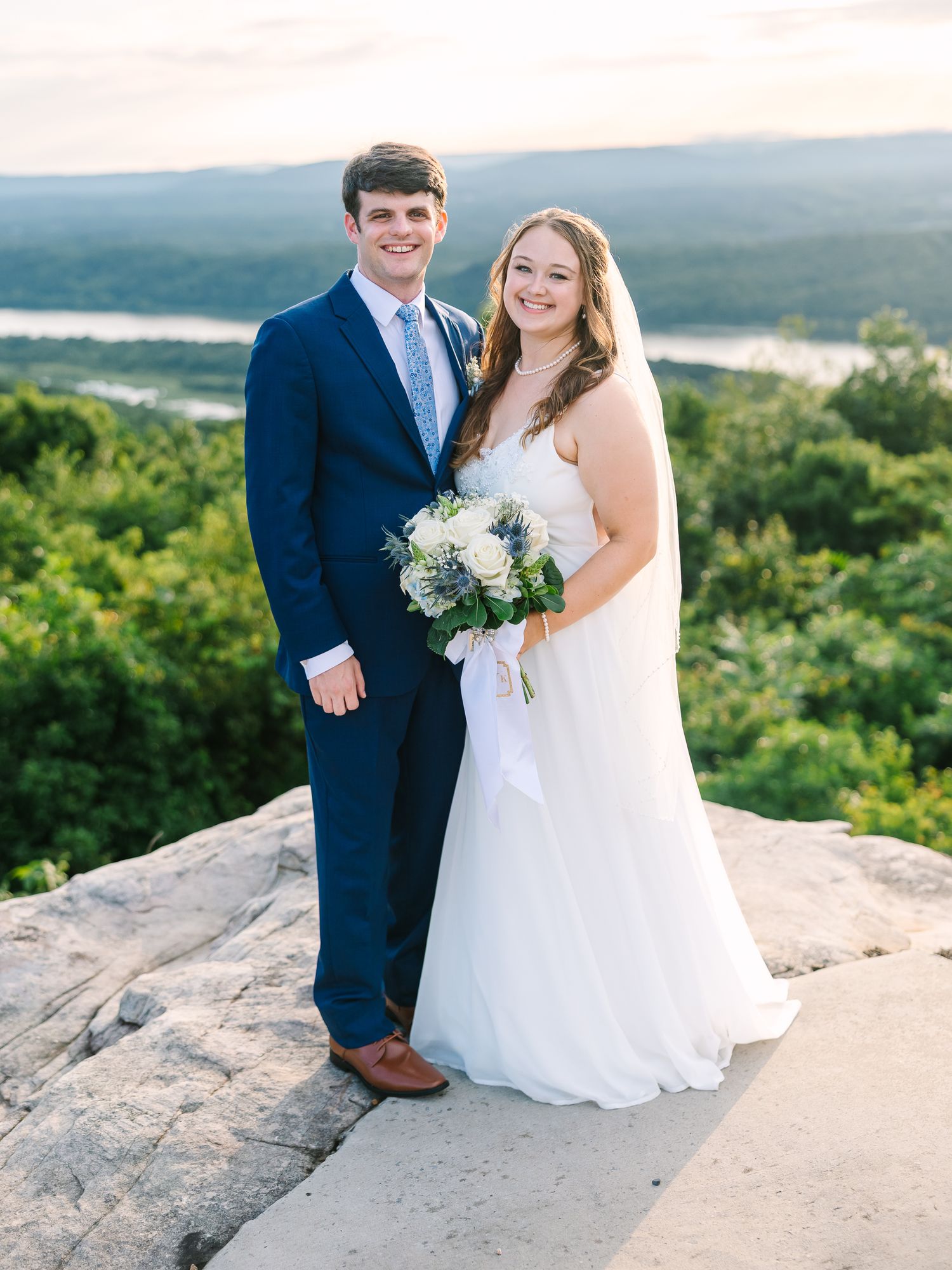 Bride and groom posing on a rocky overlook with lush green hills and a river in the background at sunset.