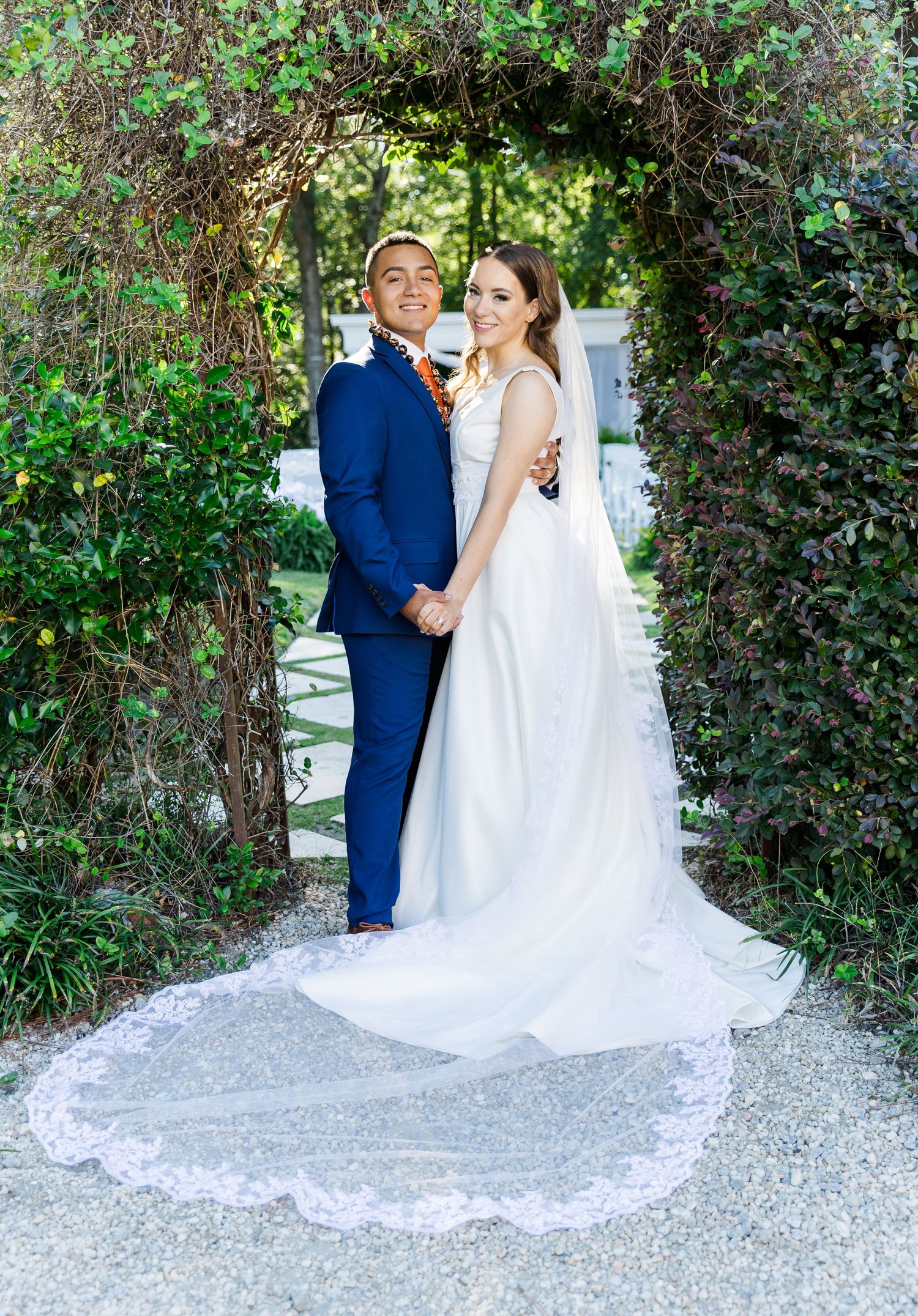 Bride and groom pose under a lush garden archway, she in a white gown with lace veil, he in a navy suit.