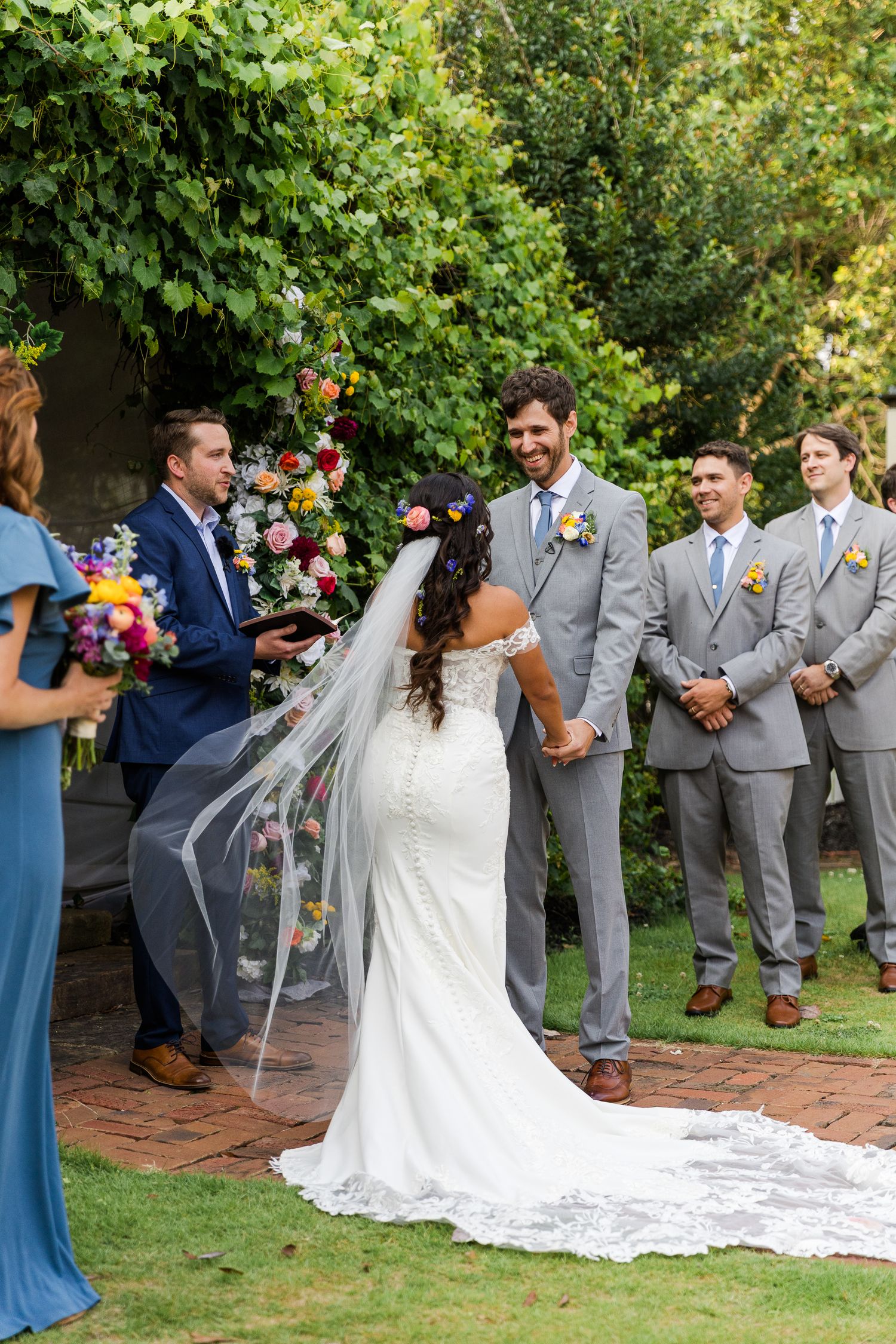 Wedding party in grey suits and blue dresses gathered outdoors for ceremony photos.