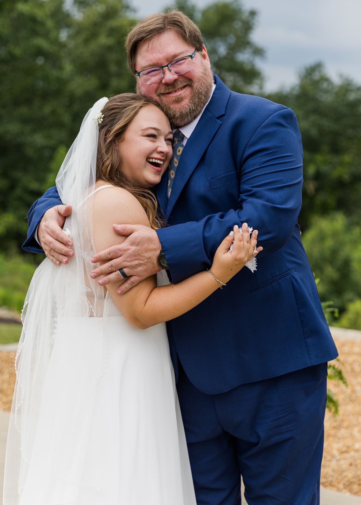 Joyful bride in white gown and veil hugging a man in a navy blue suit at an outdoor wedding ceremony.