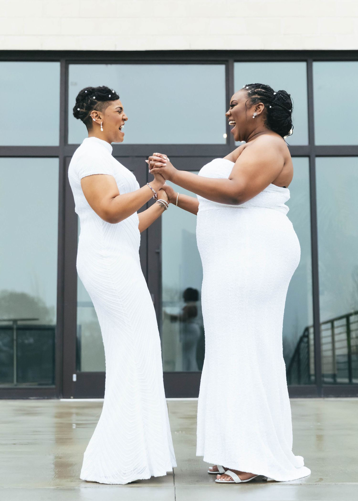 Two women in white gowns holding hands and smiling at each other outdoors, celebrating their wedding day.