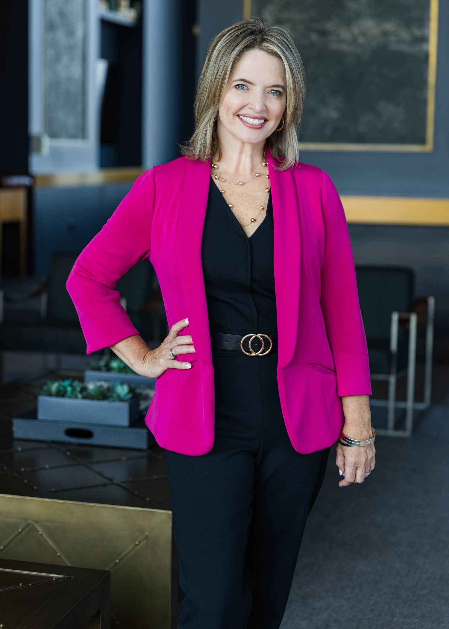 Professional woman in a bright pink blazer and black outfit posing in a modern office setting.